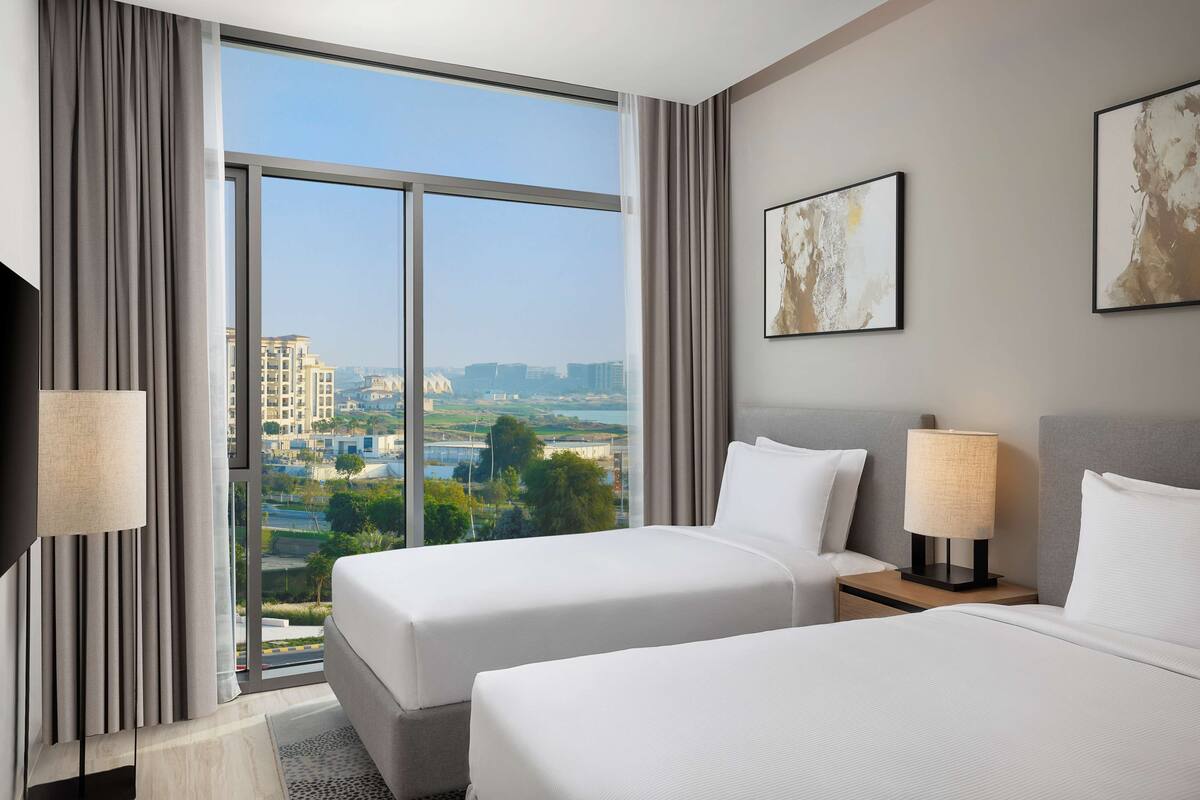 A hotel room with two beds, a large window with a city view, a flat-screen TV, and two wall-mounted lamps.