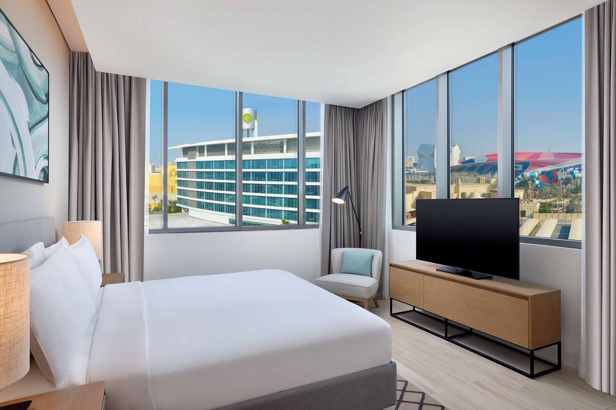 A hotel room with a large bed, a TV on a wooden console, a chair, and a view of a cityscape through large windows.