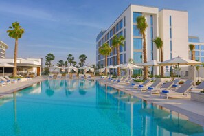 Una piscina con sillas de descanso y sombrillas, rodeada de palmeras y un edificio moderno.