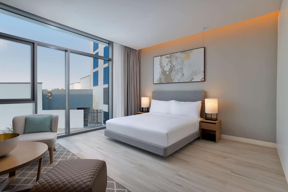 A modern hotel room with a large bed, two bedside lamps, a sofa, a small table, and a large window offering a view of the cityscape.