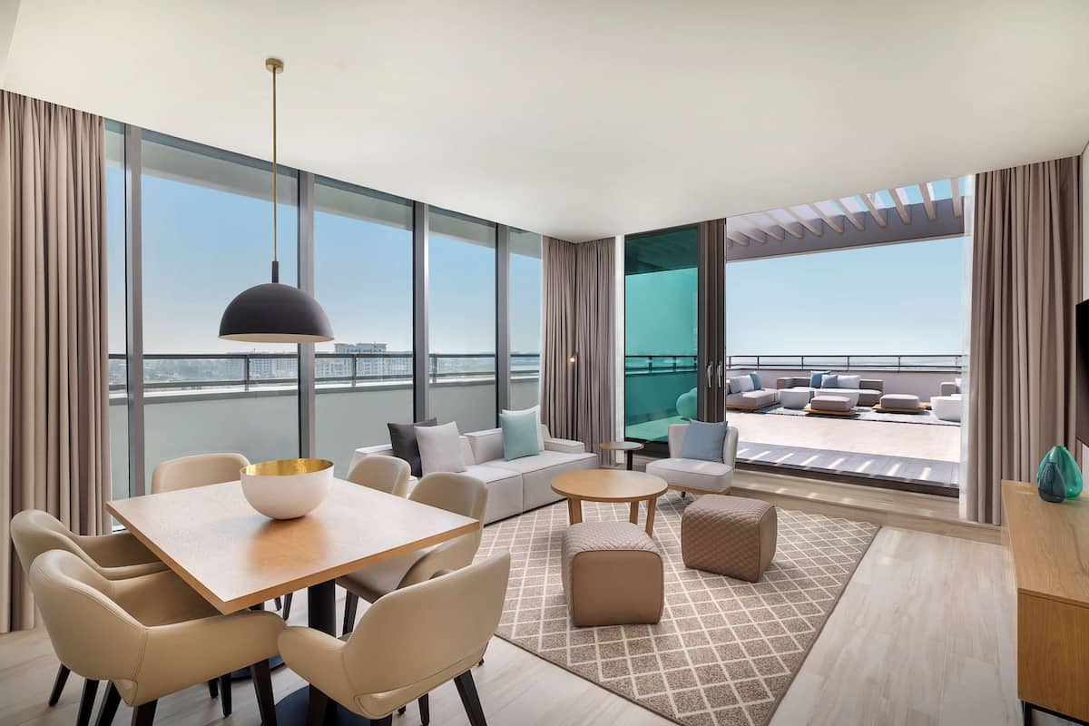 A modern hotel room with a large balcony, a dining area, and a seating area with a view of the city.