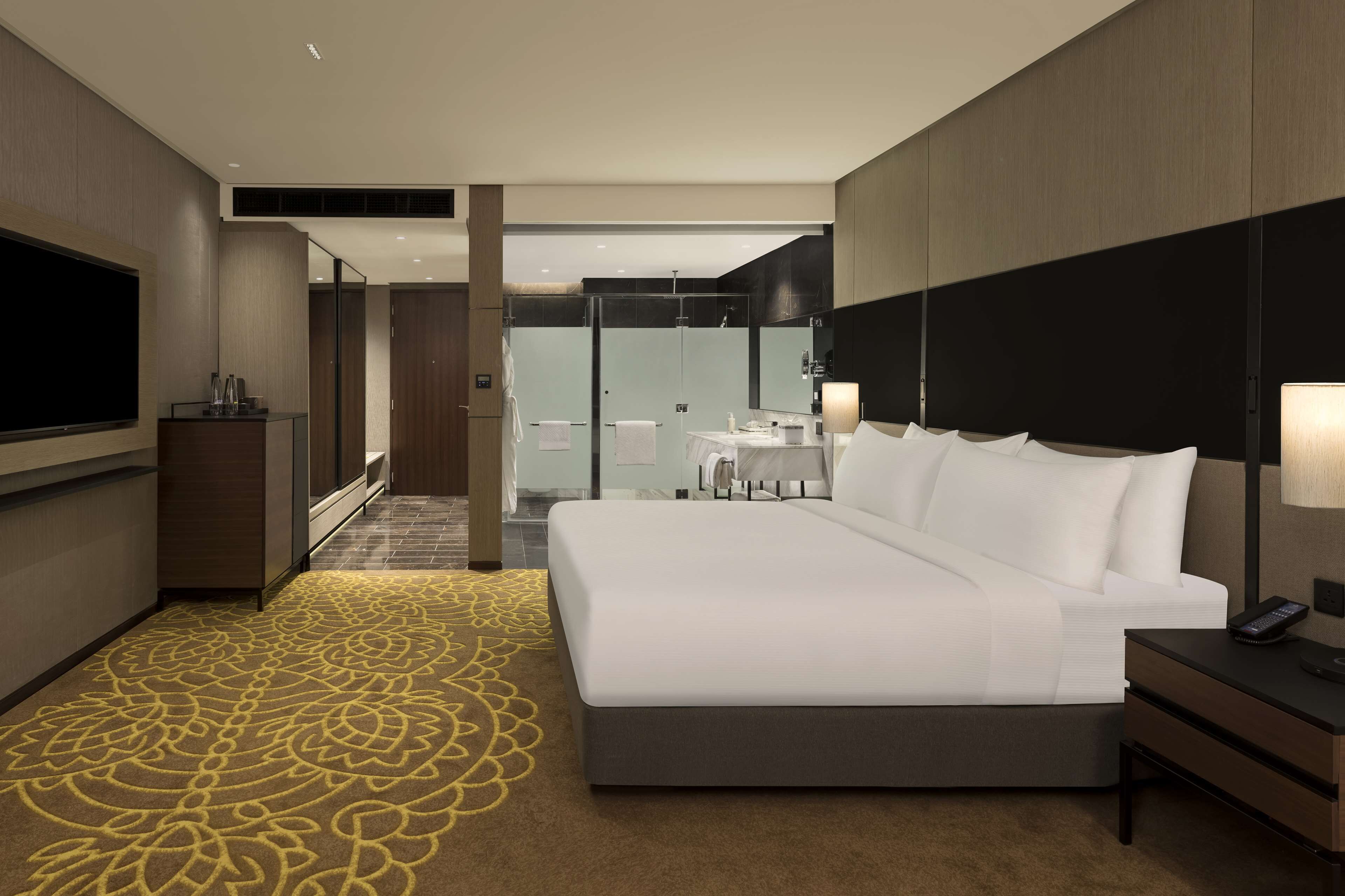 A modern hotel room with a large bed, a TV, a desk with a lamp, and a bathroom visible through a glass door.