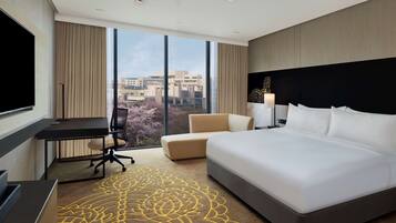 A hotel room with a large bed, a desk, a chair, a TV, and a view of the city through large windows.