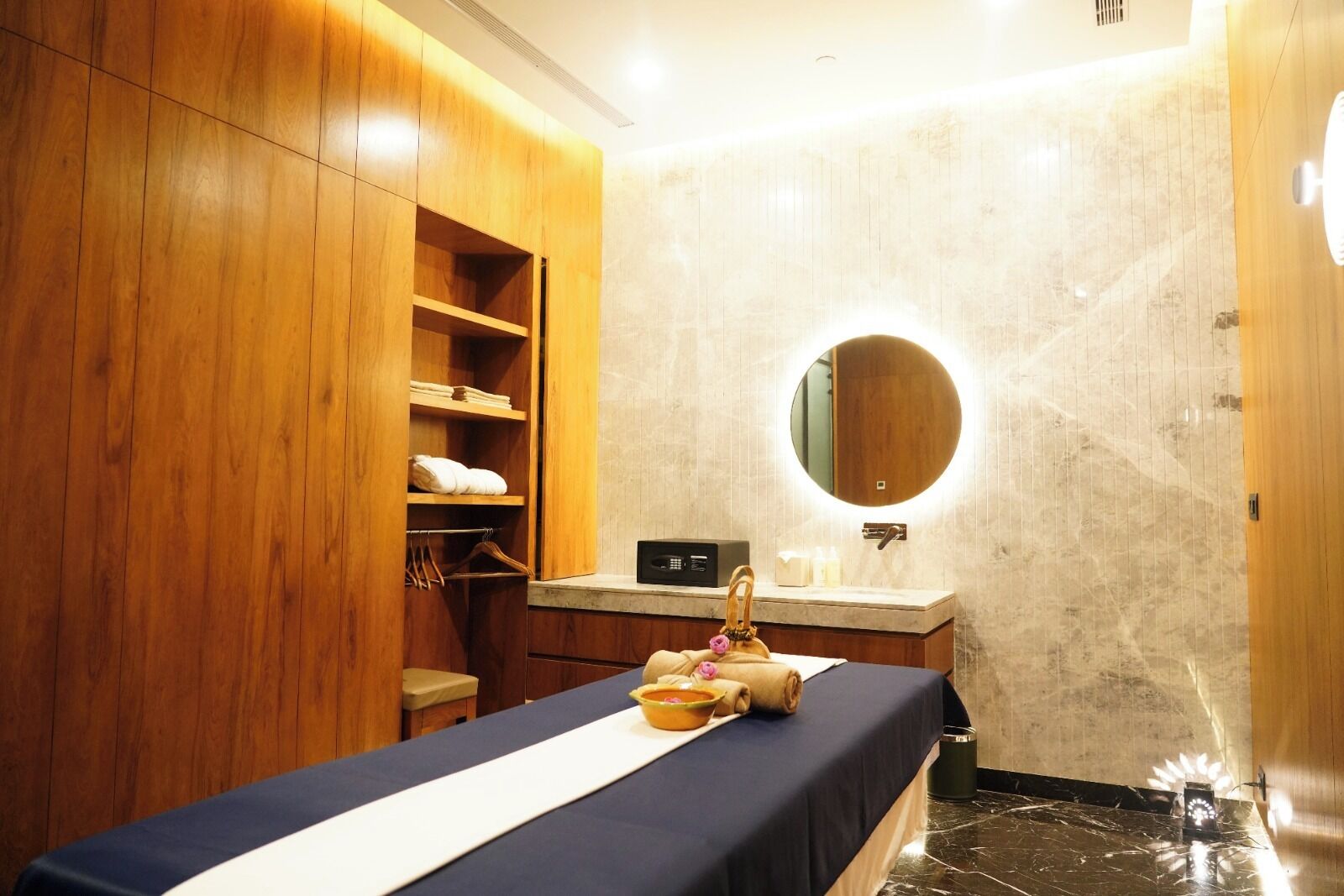 A hotel bathroom with a wooden wardrobe, a round mirror, a sink, and a massage table.