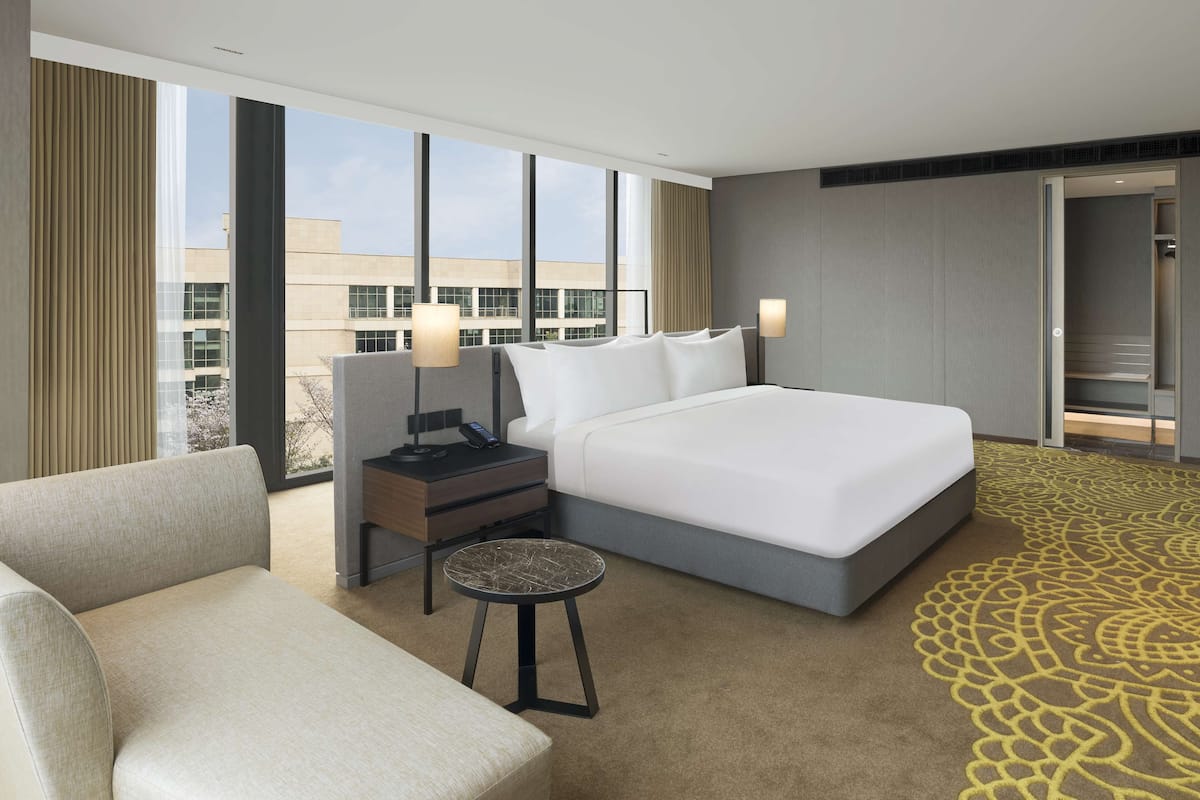 Une chambre d’hôtel moderne avec un grand lit, une table de chevet avec une lampe, une petite table ronde et une vue sur la ville grâce à de grandes fenêtres.