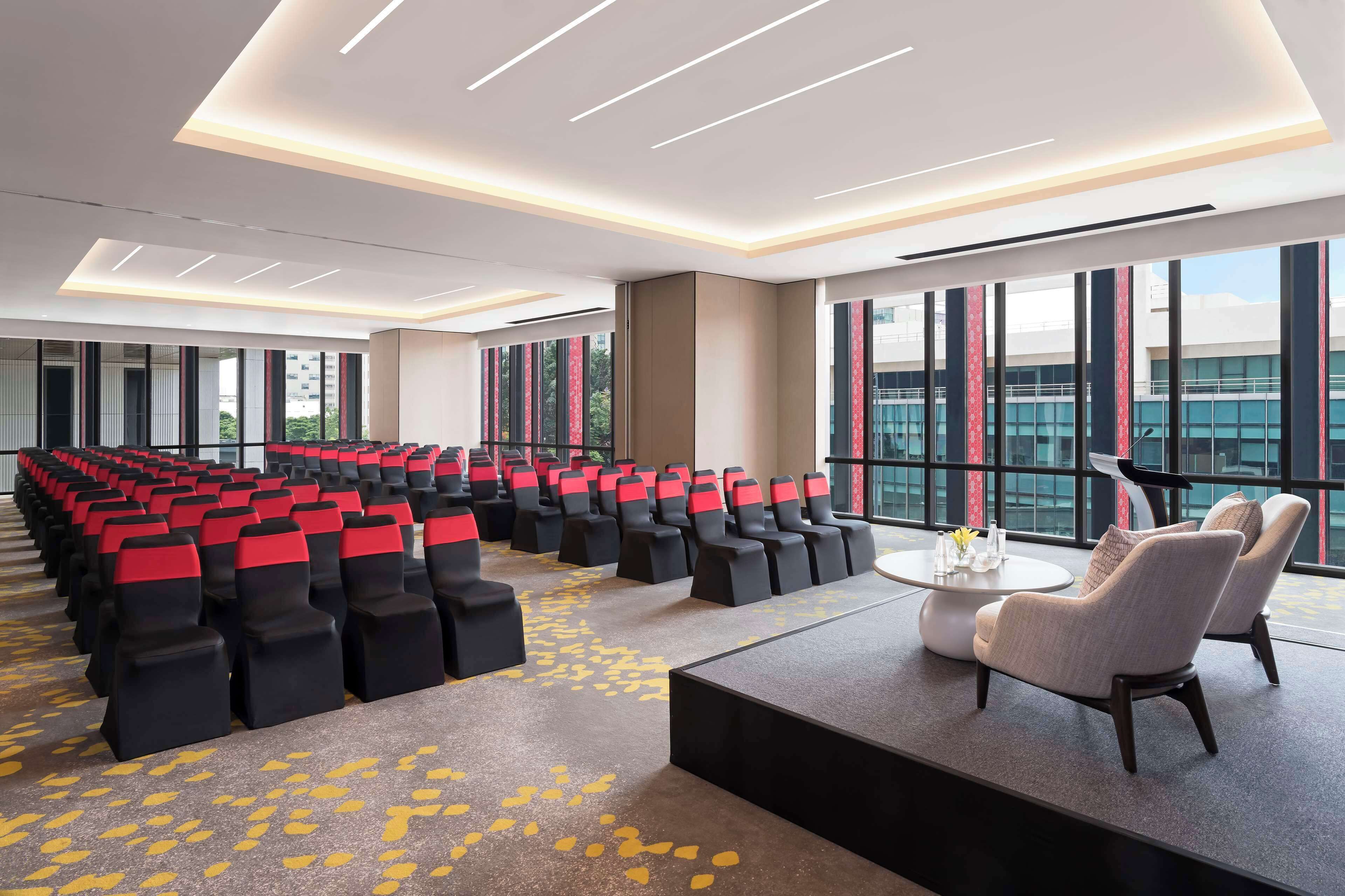 A conference room with rows of chairs, a stage with a podium, and large windows offering a view of the city.
