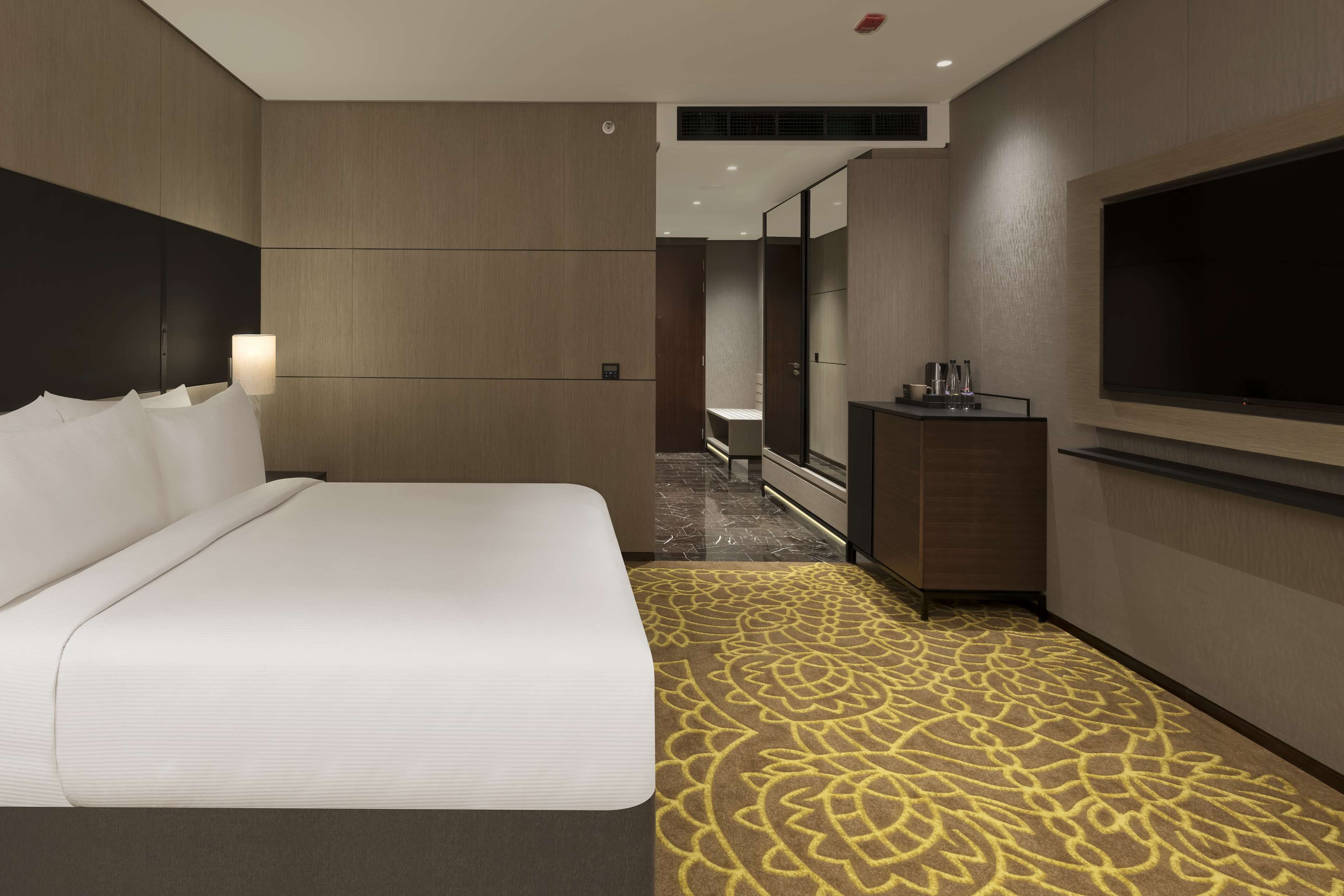 A hotel room with a large bed, a flat-screen TV mounted on the wall, a small desk with a mirror, and a patterned carpet.