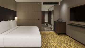A hotel room with a large bed, a flat-screen TV mounted on the wall, a small desk with a mirror, and a patterned carpet.