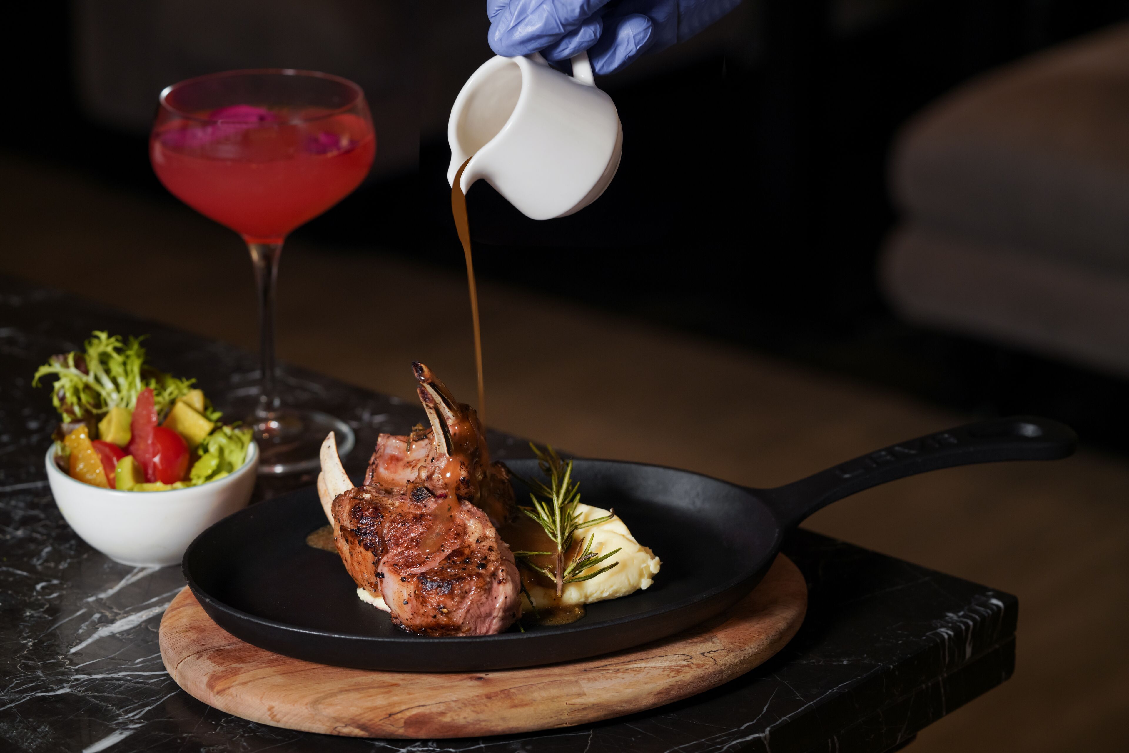A dish of grilled lamb chops with rosemary and mashed potatoes on a black skillet, a bowl of salad, and a red cocktail on a marble table.