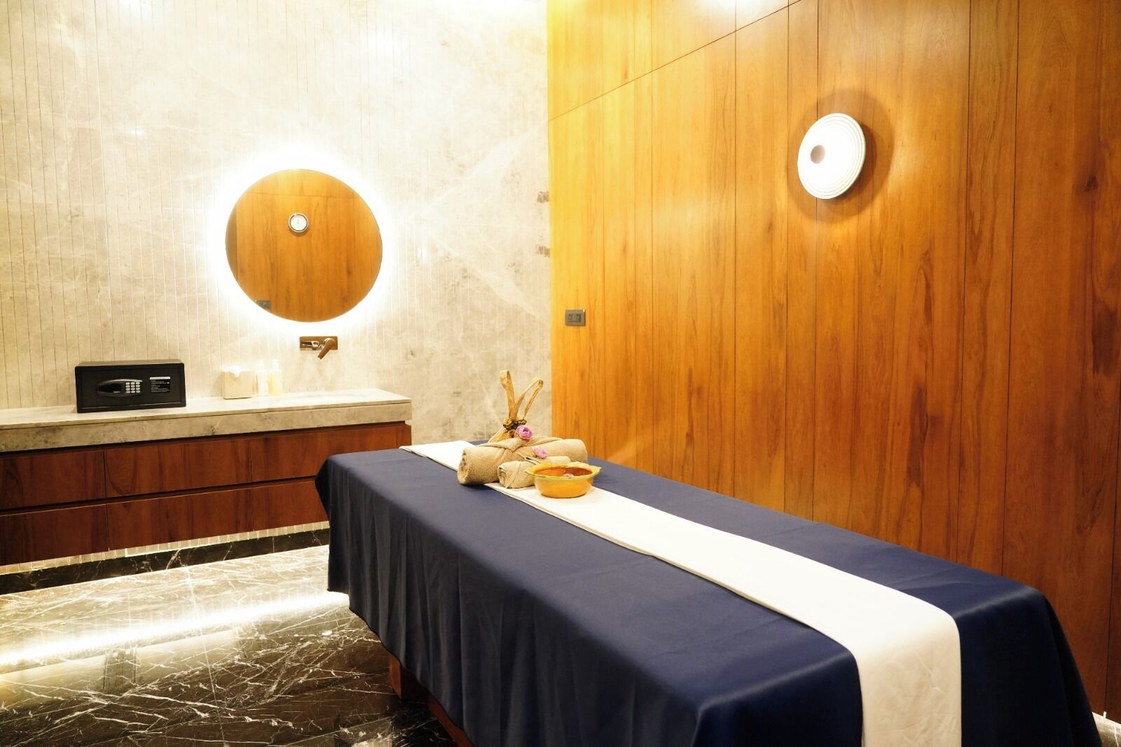 A room with a wooden wall, a round mirror, a wooden cabinet, a massage table with towels and a bowl, and a marble floor.