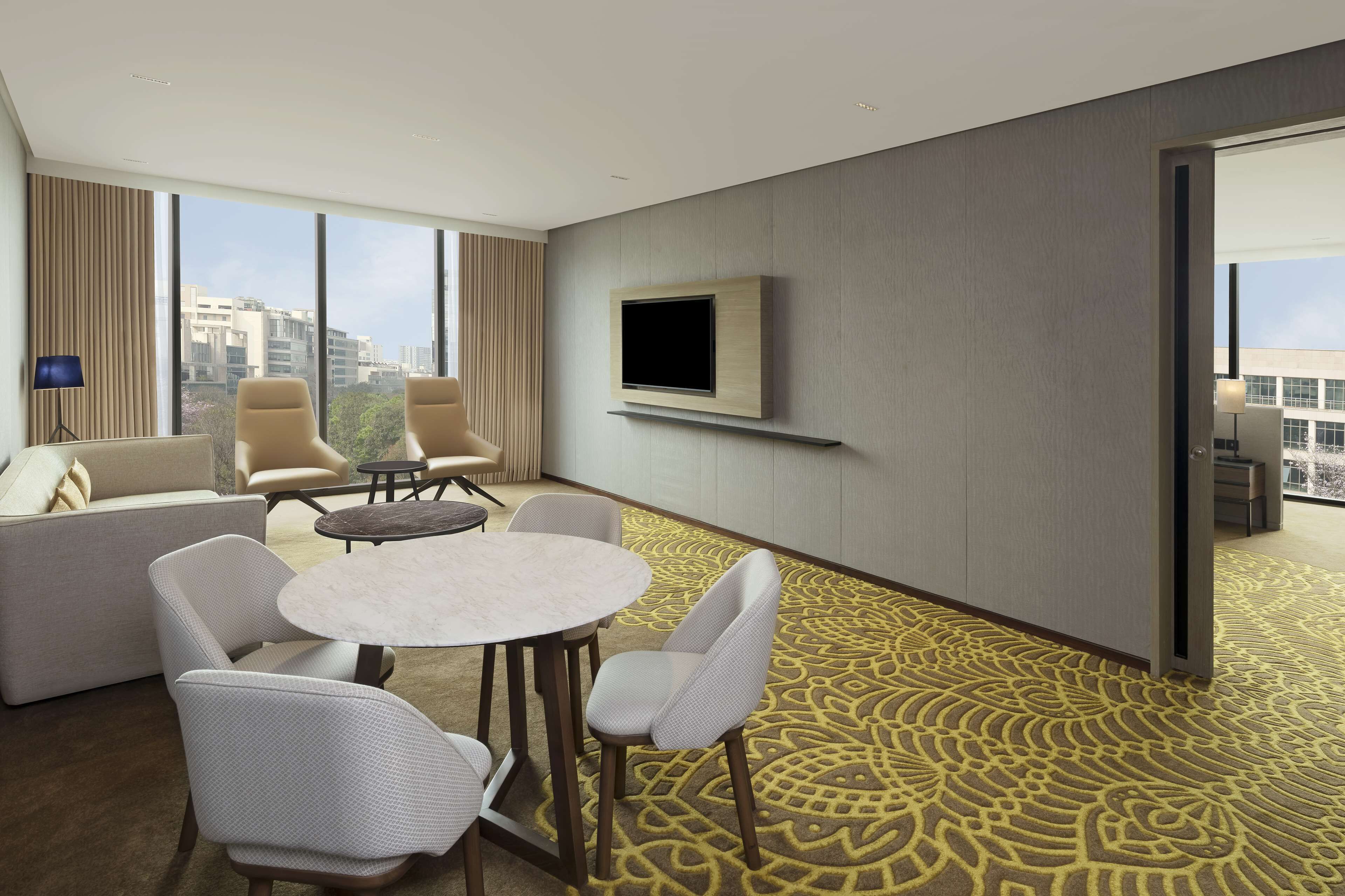 A modern hotel room with a large window, a flat-screen TV mounted on the wall, a sofa, two armchairs, a small round table, and a patterned carpet.