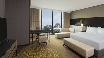 A hotel room with a large bed, a desk, a chair, and a sofa. There is a TV on the left side of the room.