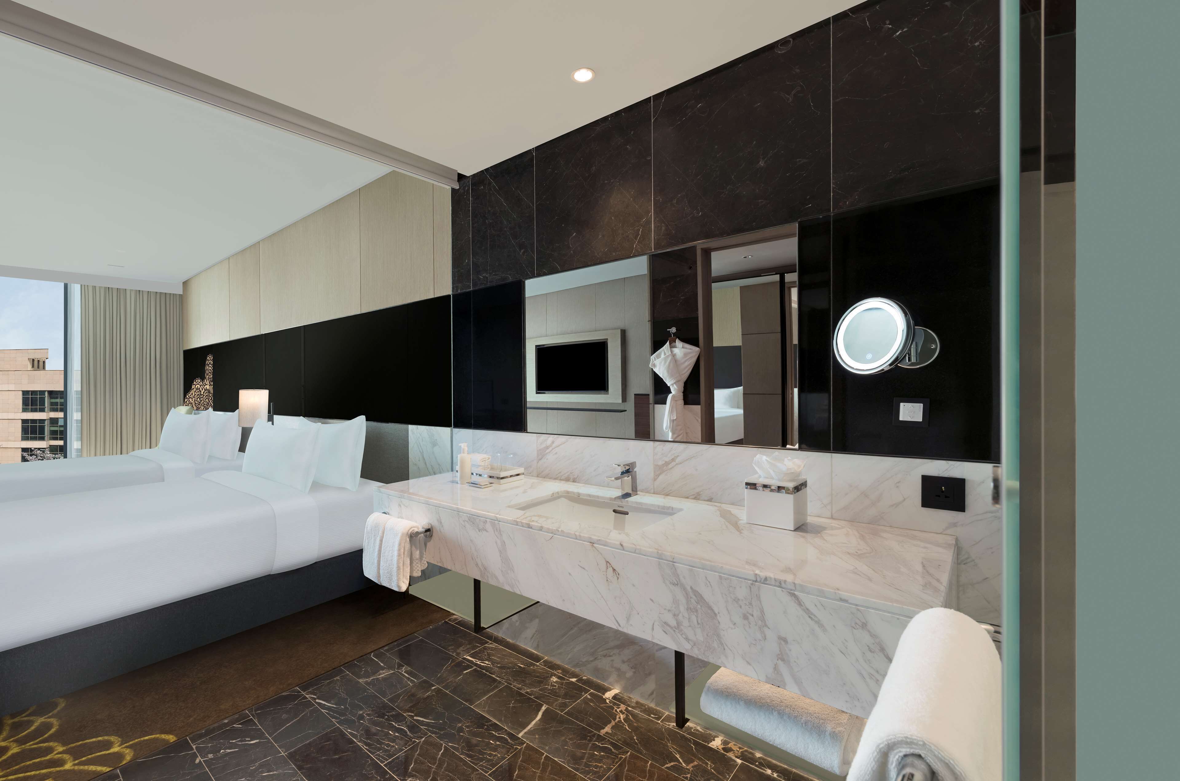 A modern hotel room with a large bed, a marble bathroom vanity, a large mirror, and a television.