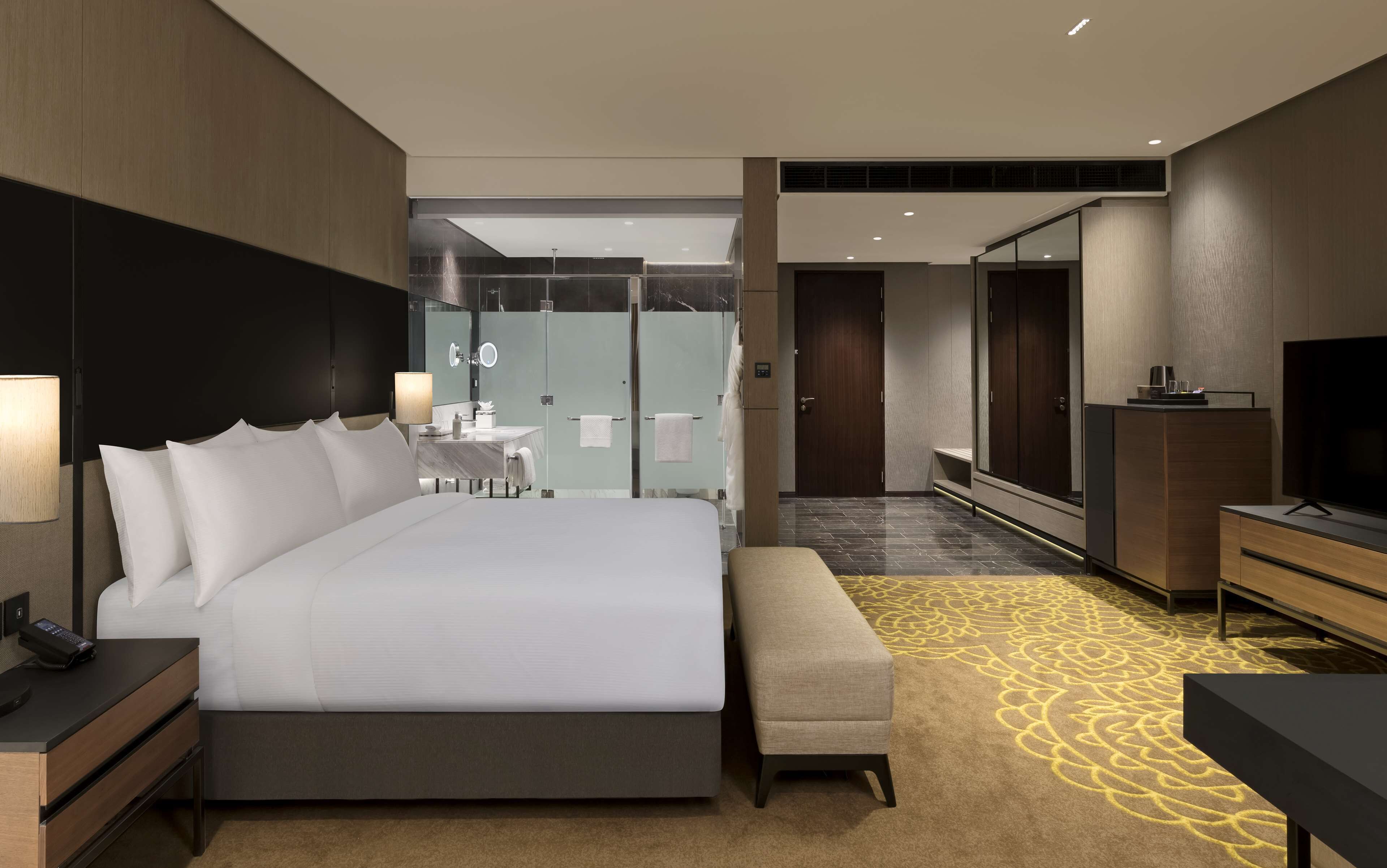 Habitación de hotel moderna con una cama grande, mesitas de noche, un escritorio, un banco, un televisor y un baño con espejo, toalleros y ducha.