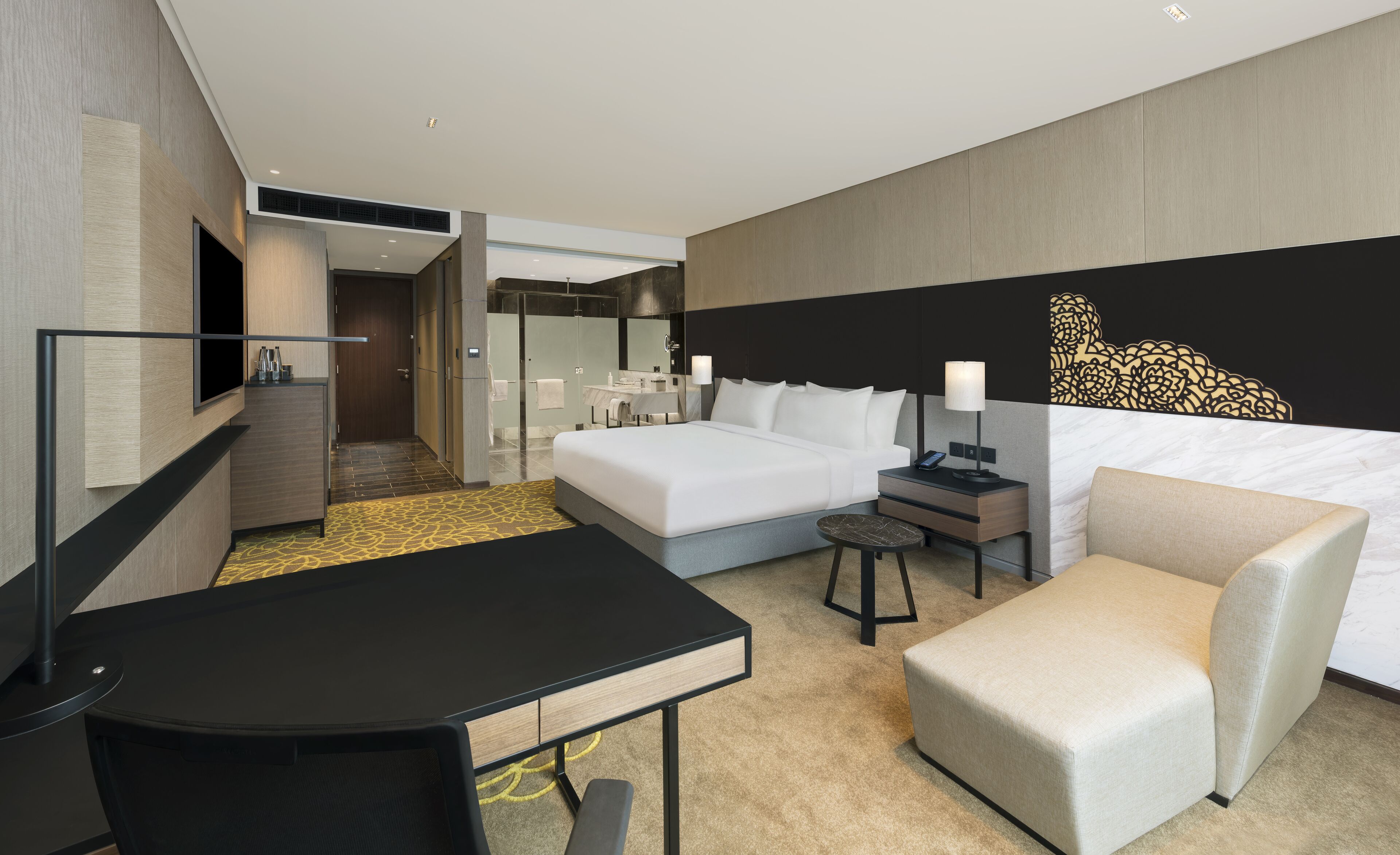 A modern hotel room with a bed, a desk, a chair, a small table, a bathroom, and a decorative wall piece.