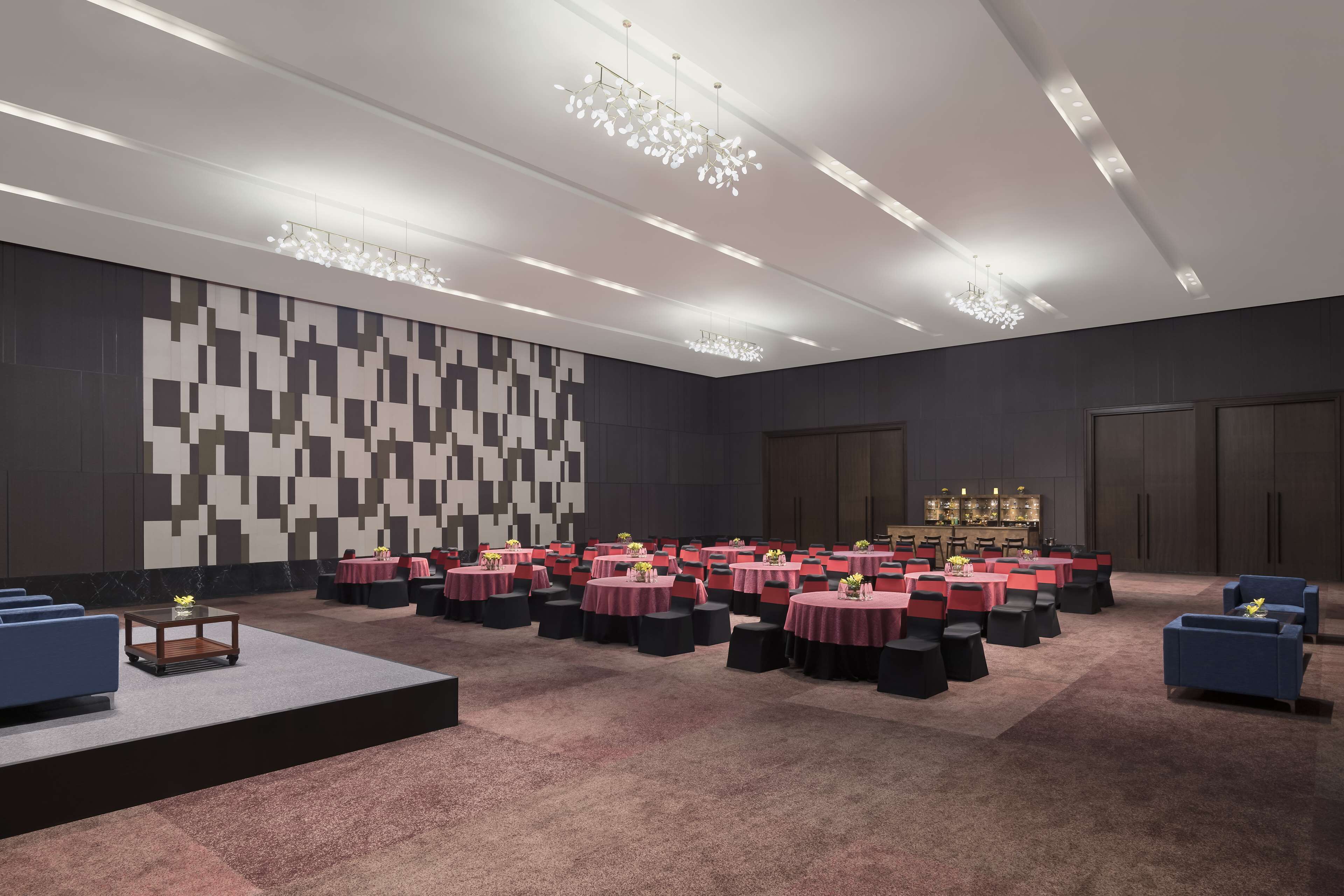 Una espaciosa sala de conferencias con varias mesas y sillas, un escenario y una pared grande con un diseño geométrico.