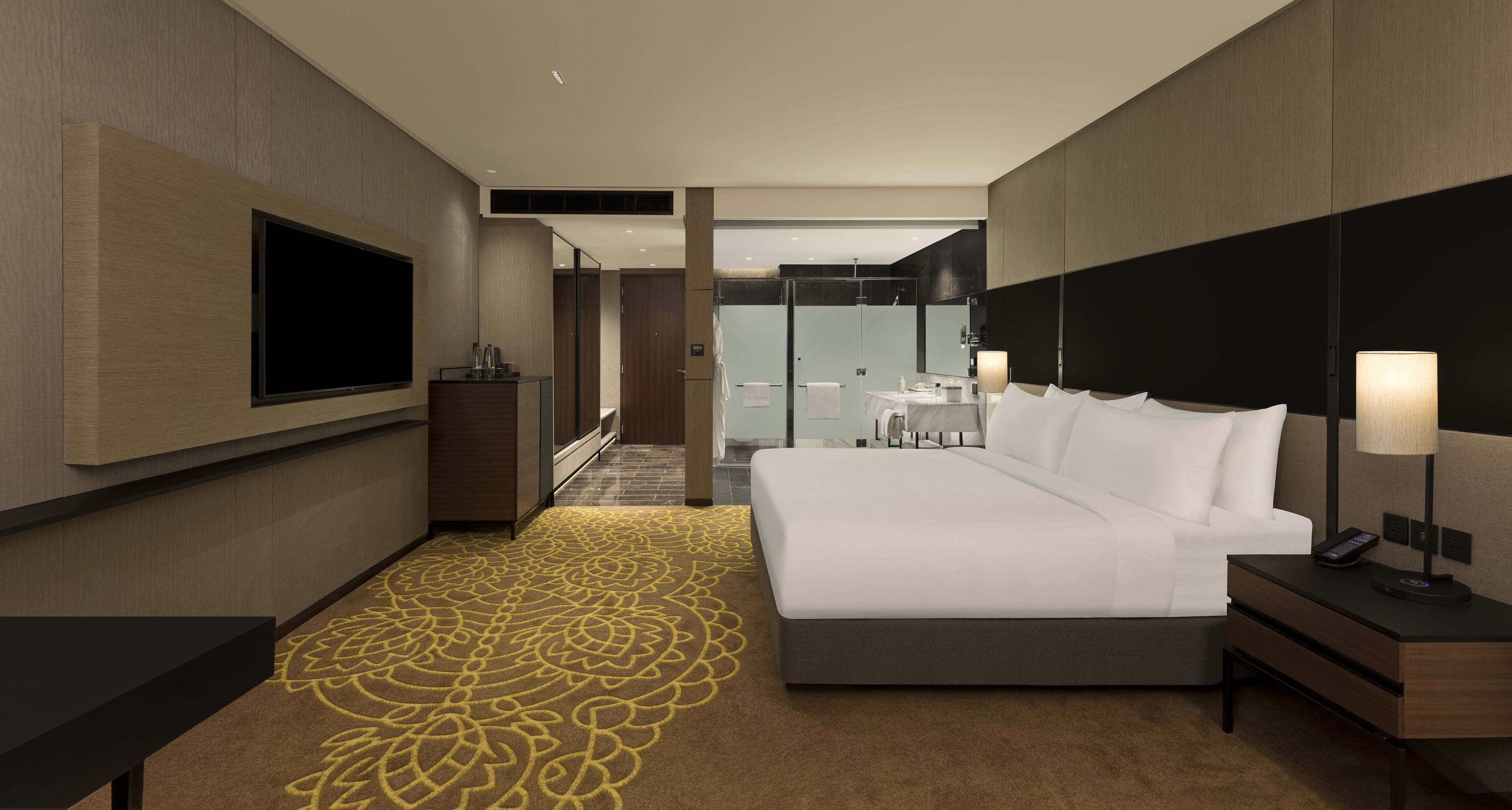 A modern hotel room with a large bed, a TV mounted on the wall, a bedside table with a lamp, and a bathroom visible through a glass door.