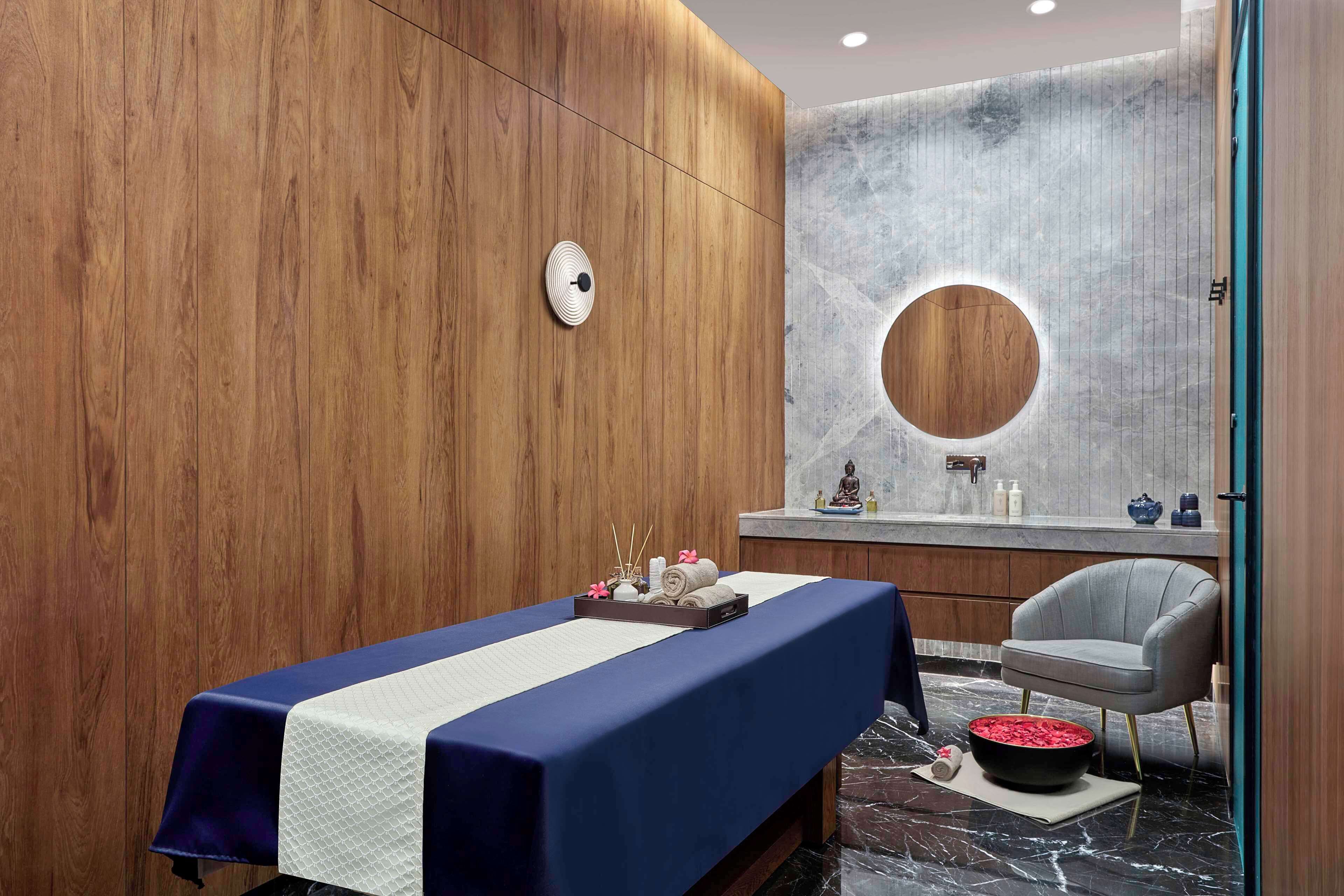 A modern spa room with a wooden wall, a massage table, a round mirror, and a gray chair.