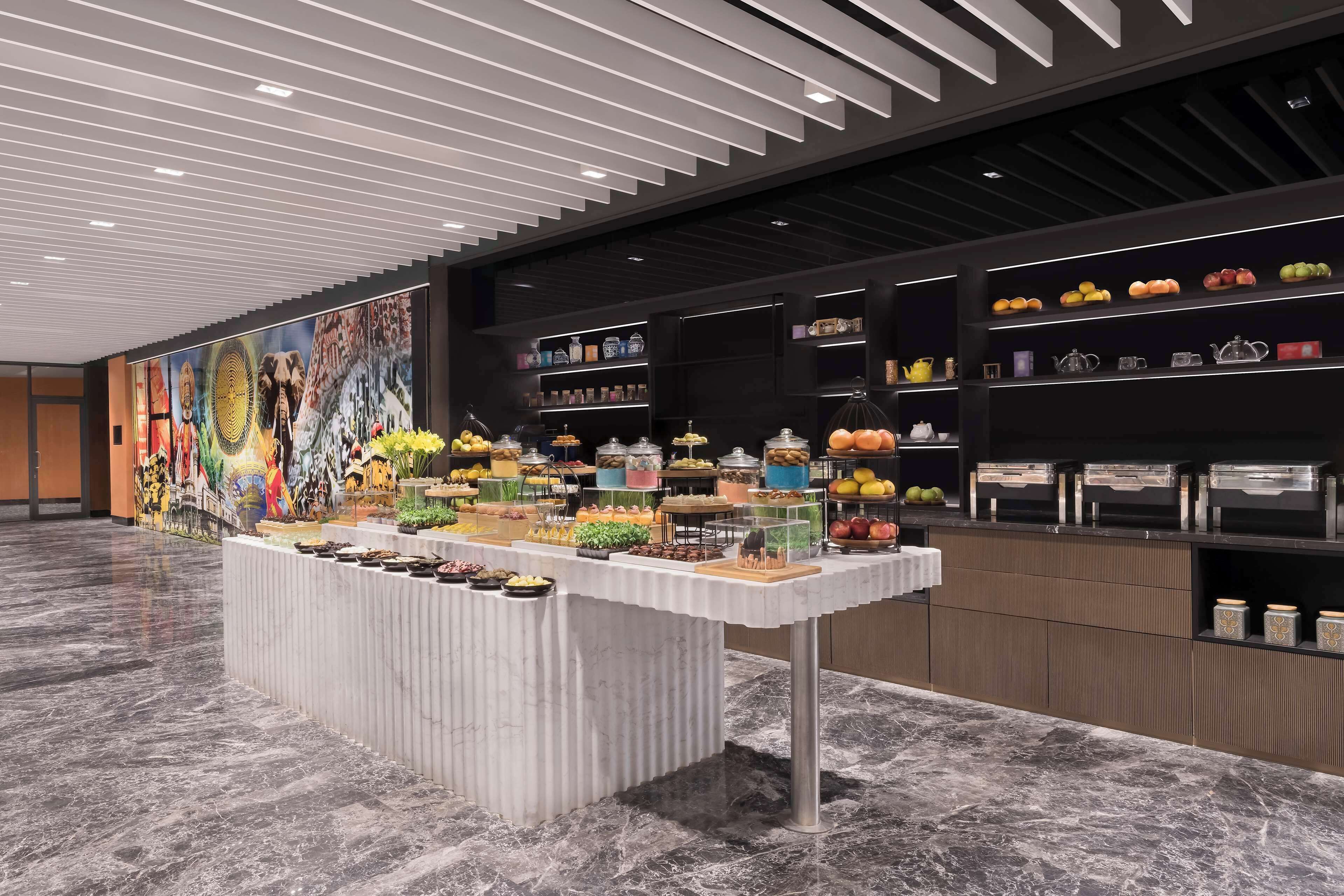 A modern hotel lobby with a marble floor, a central buffet table with various food items, and a large mural on the wall.