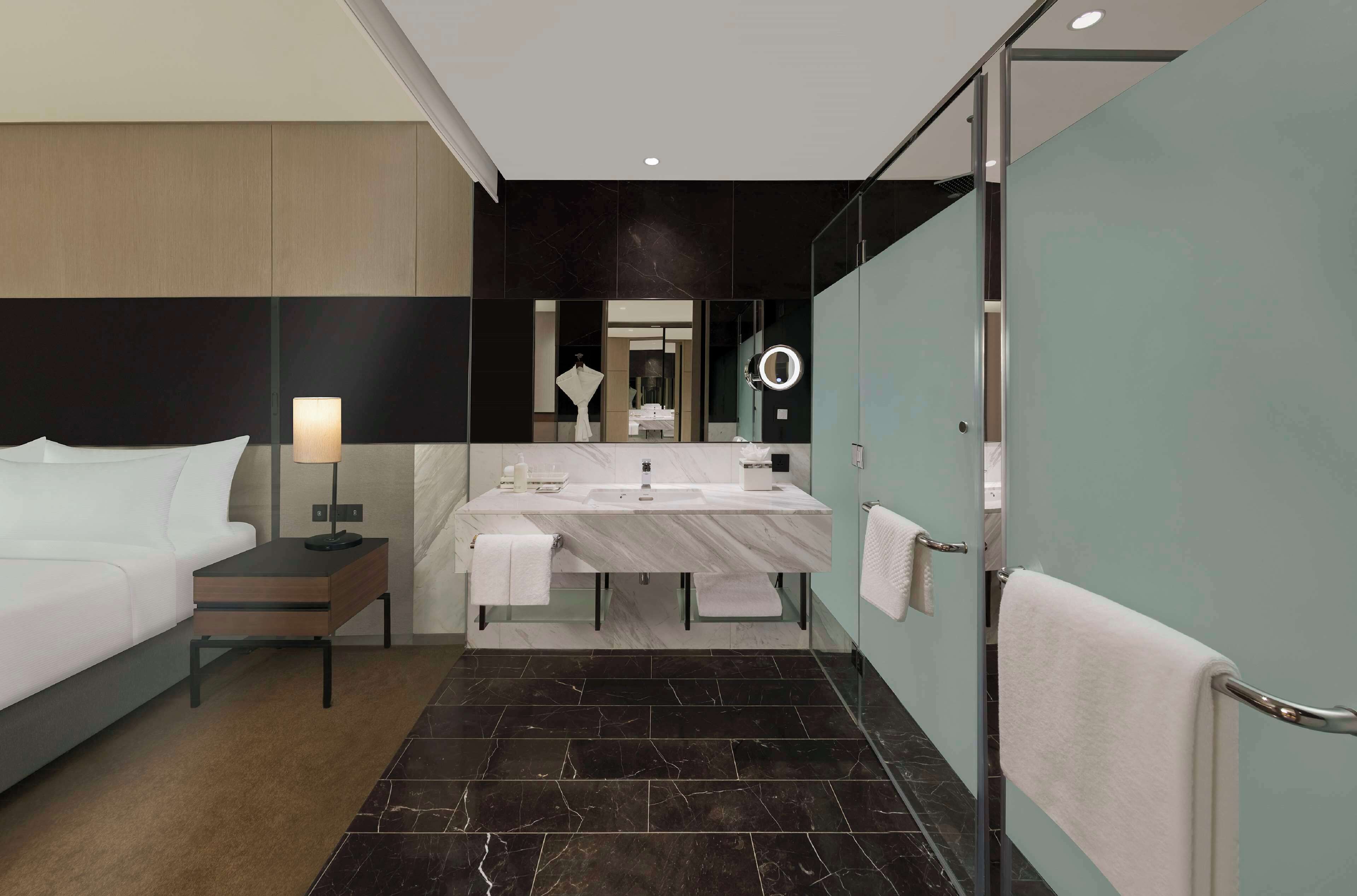 A modern hotel room with a large bed, a bedside table with a lamp, a bathroom with a large mirror, a round light, and a towel rack.