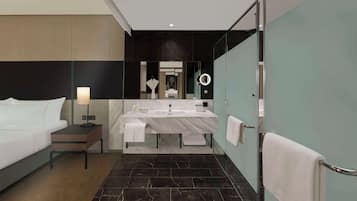 Una habitación de hotel moderna con una cama grande, una mesita de noche con lámpara, un baño con un espejo amplio, una luz redonda y un perchero para toallas.