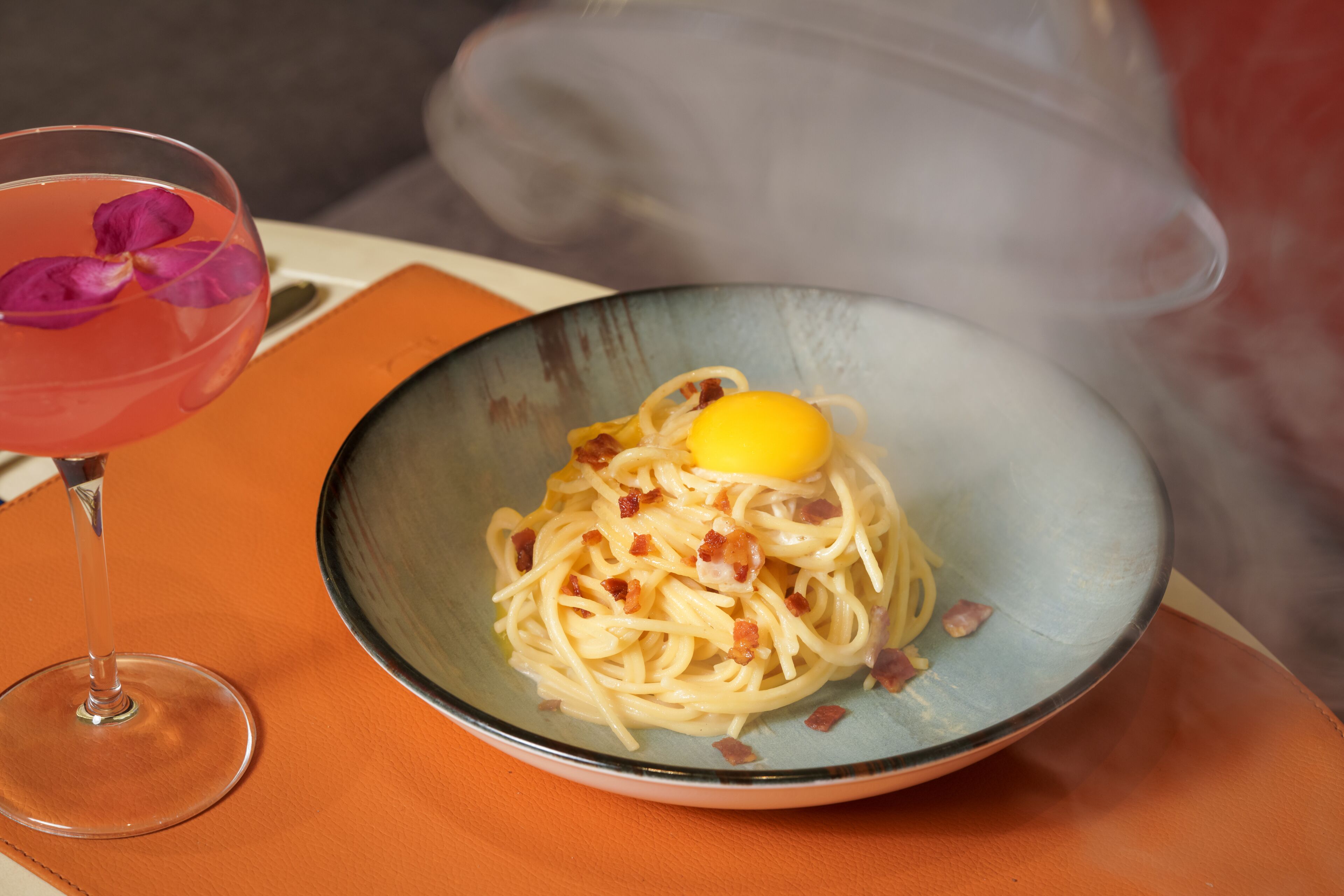 A bowl of spaghetti with bacon and a lemon wedge on an orange table.