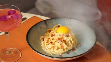 A bowl of spaghetti with bacon and a lemon wedge on an orange table.