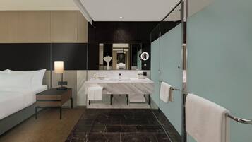 A modern hotel room with a bed, a bedside table with a lamp, a large mirror, a bathroom with a sink and a toilet, and a towel rack.