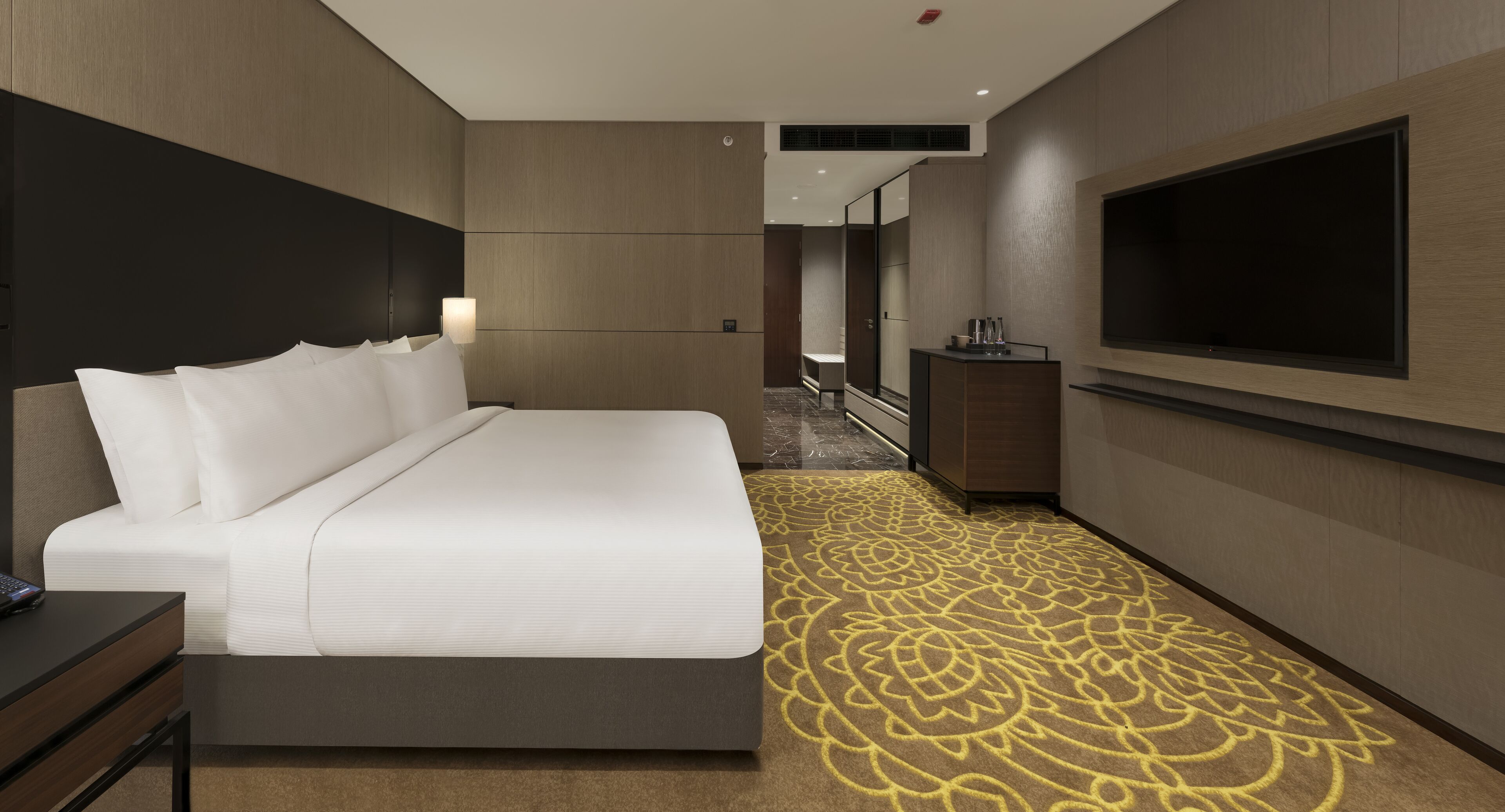 A modern hotel room with a large bed, a flat-screen TV, a desk, and a patterned carpet.