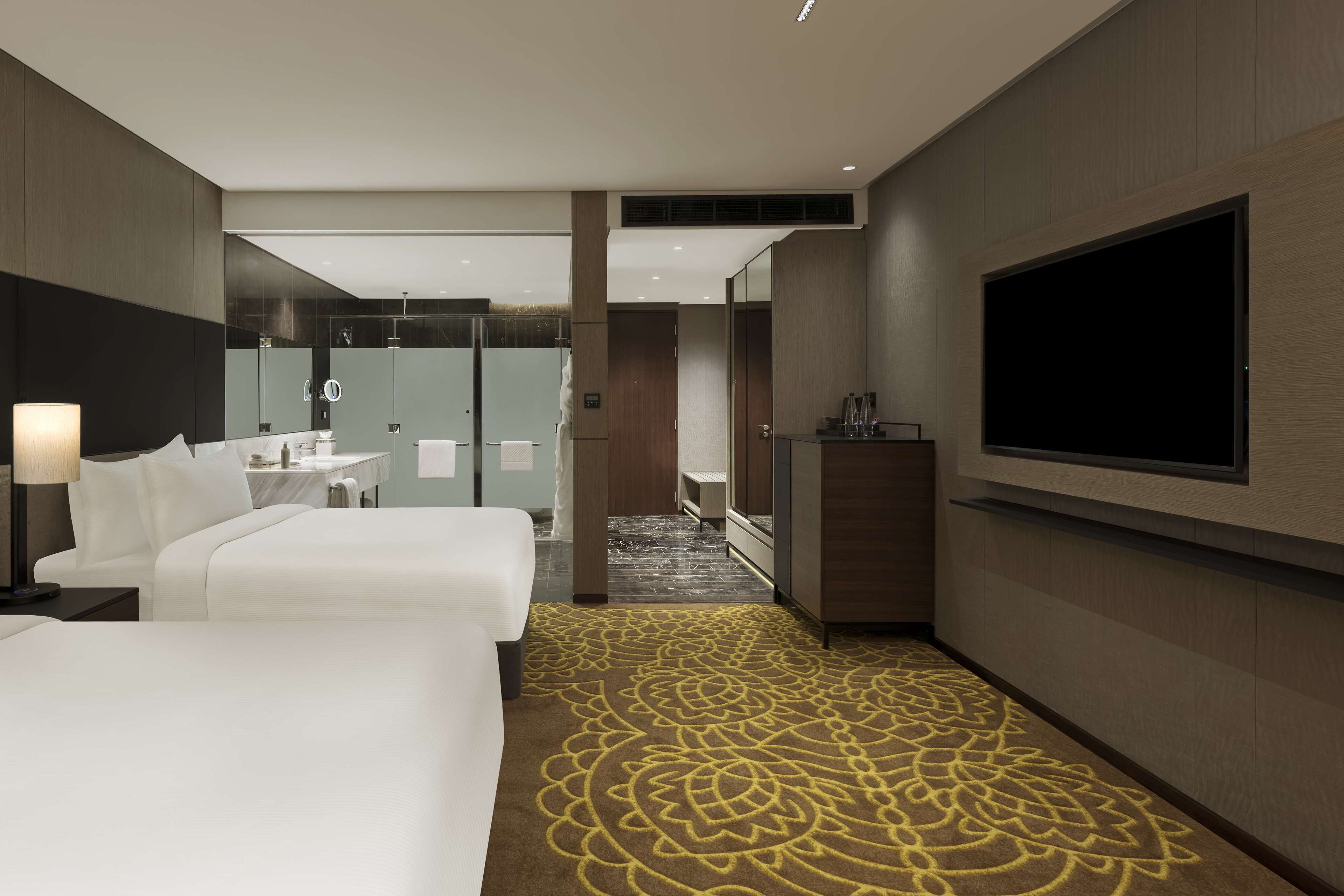 A hotel room with two beds, a TV, a bathroom with a mirror, and a patterned carpet.