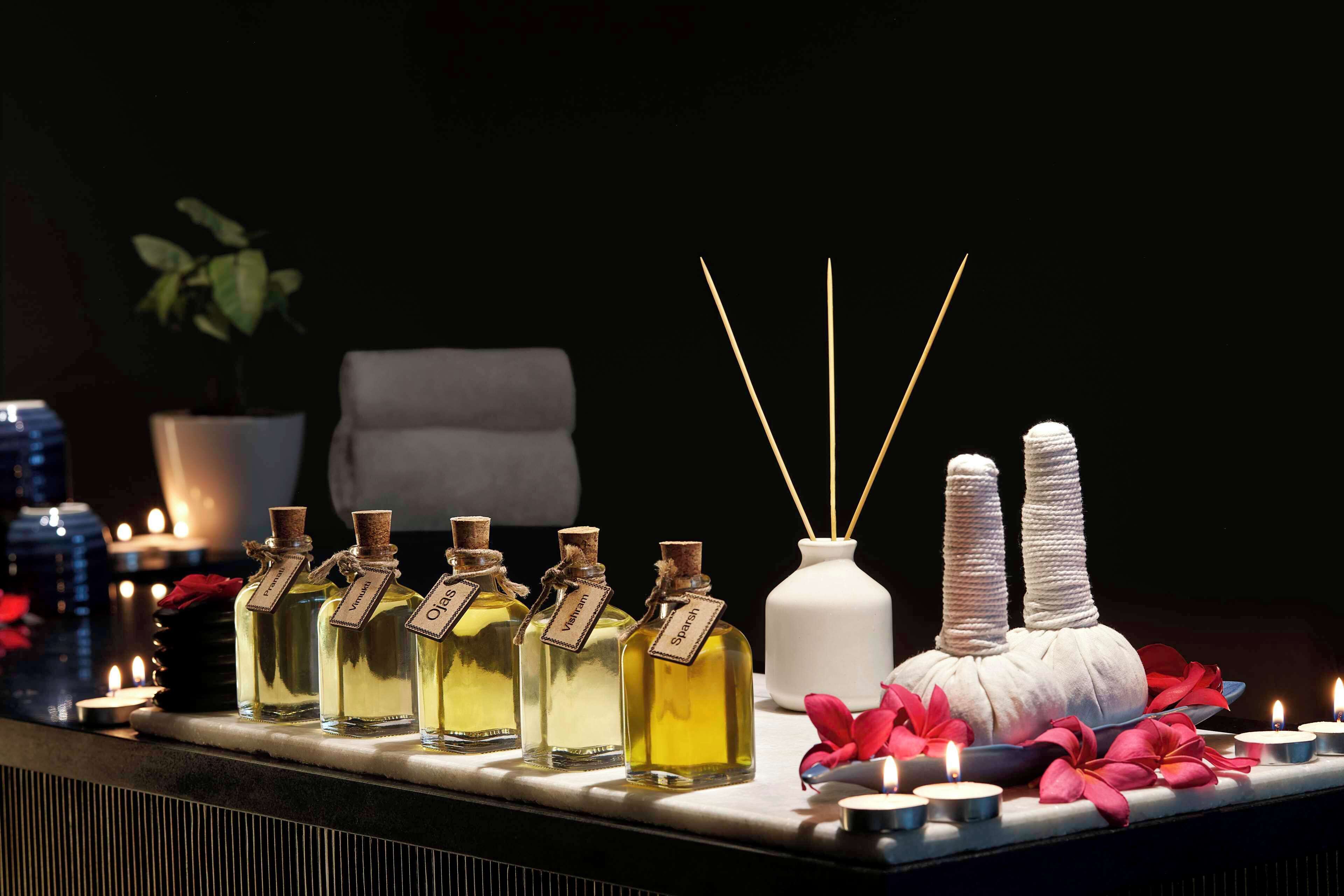 Una fila de frascos perfumados con etiquetas, un jarrón blanco con ramitas de caña y velas sobre una mesa.