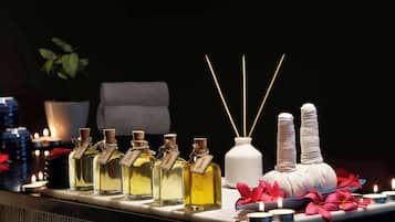 Una fila de frascos perfumados con etiquetas, un jarrón blanco con ramitas de caña y velas sobre una mesa.