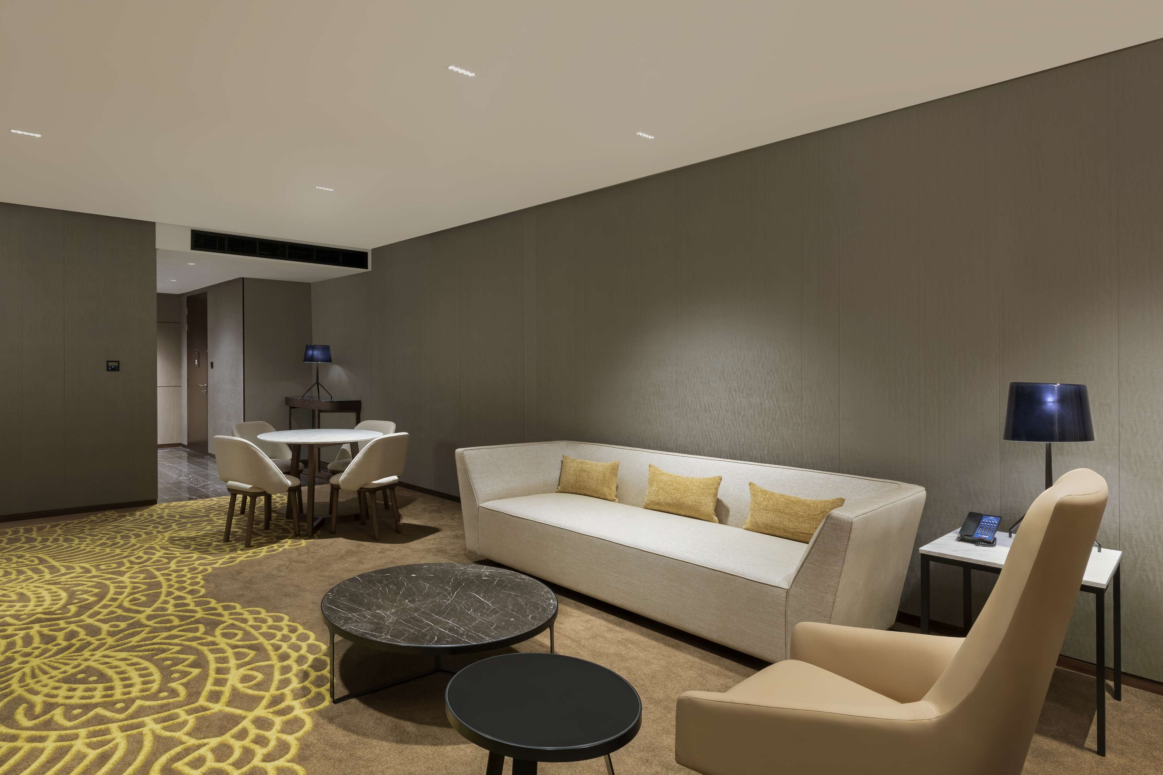 A modern hotel room with a sofa, two armchairs, a small table, a coffee table, and a patterned carpet.