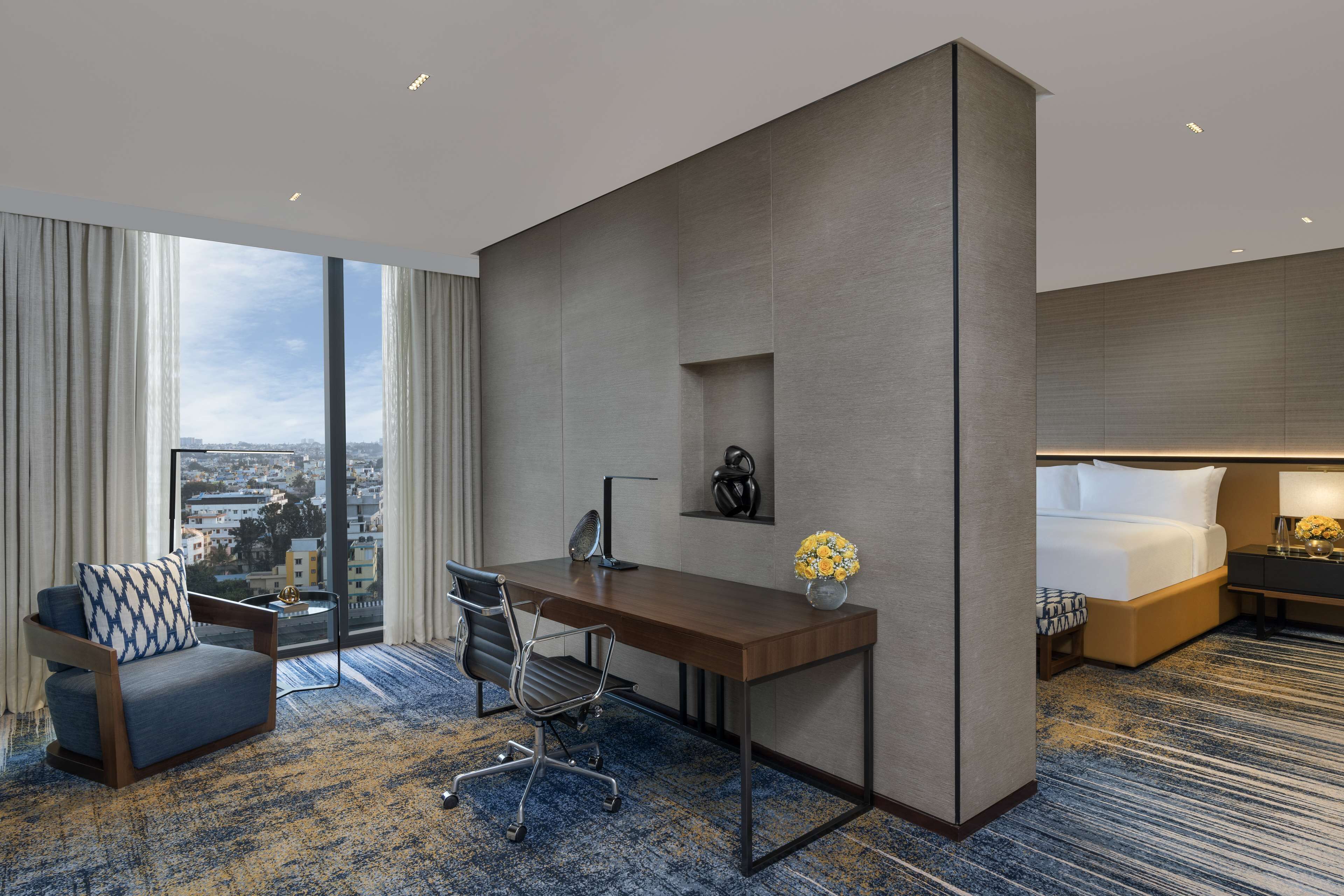 A hotel room with a large window, a desk, a chair, a bed, and a nightstand.