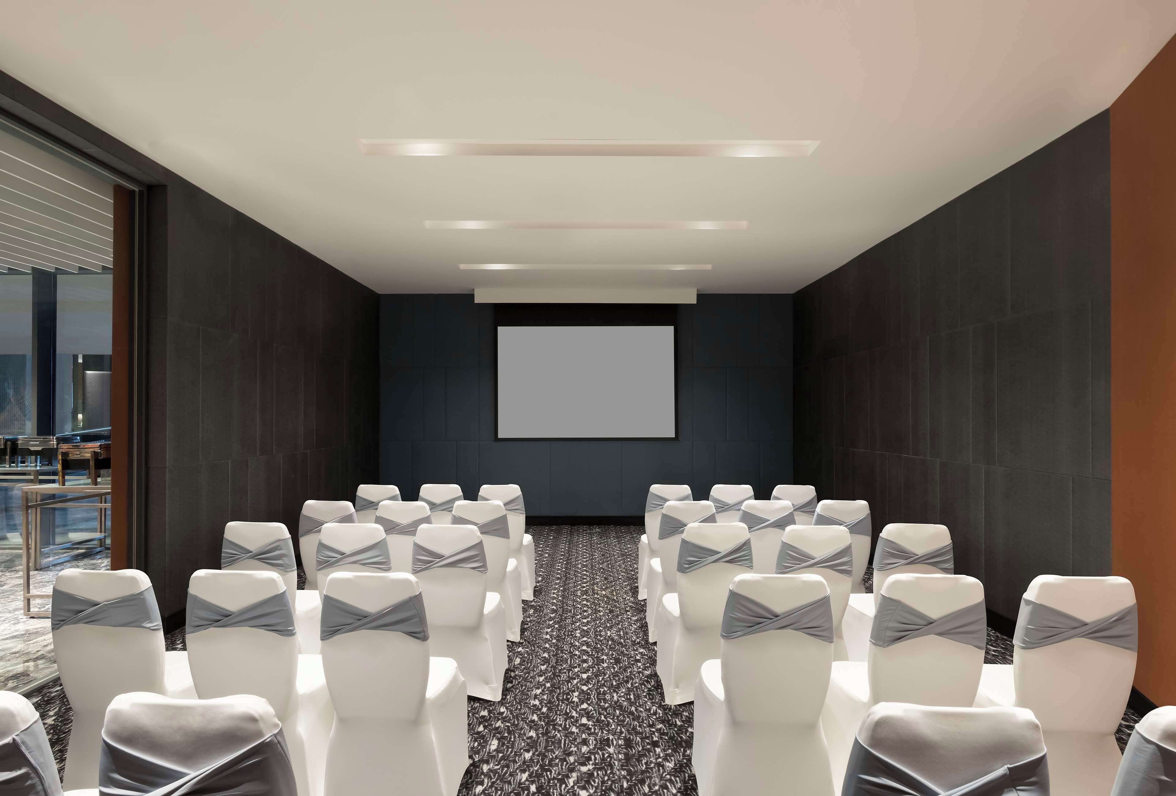 A conference room with rows of white chairs with grey sashes, a projector screen, and a dark tiled wall.