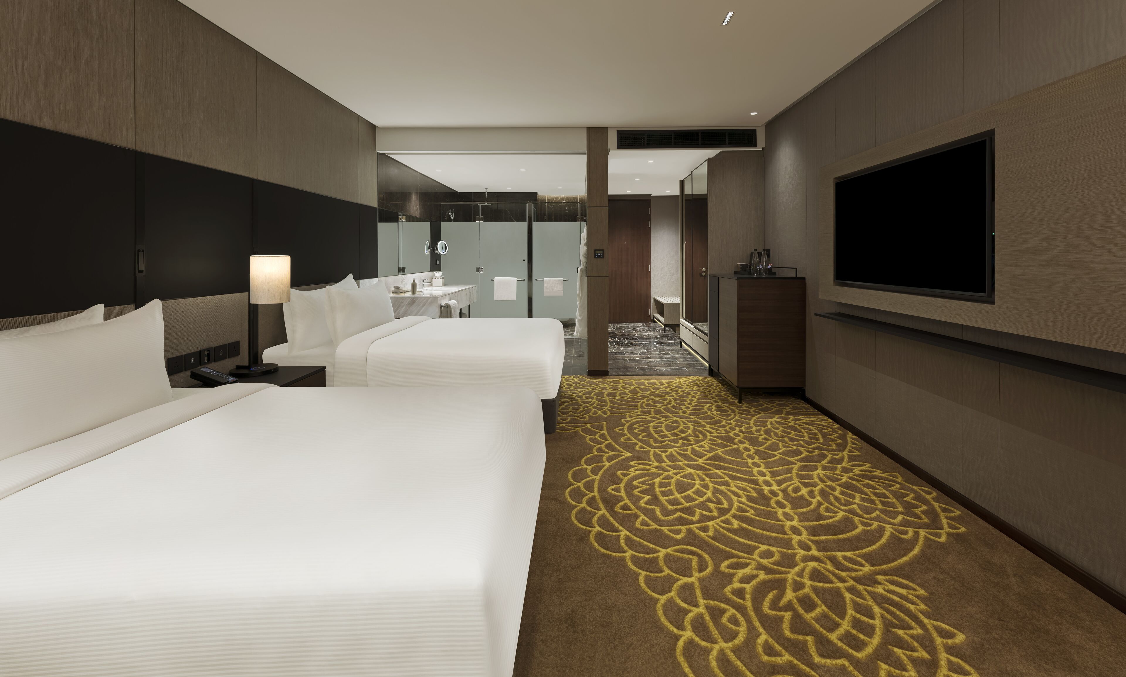 A hotel room with two beds, a TV, a desk, and a bathroom visible in the background.