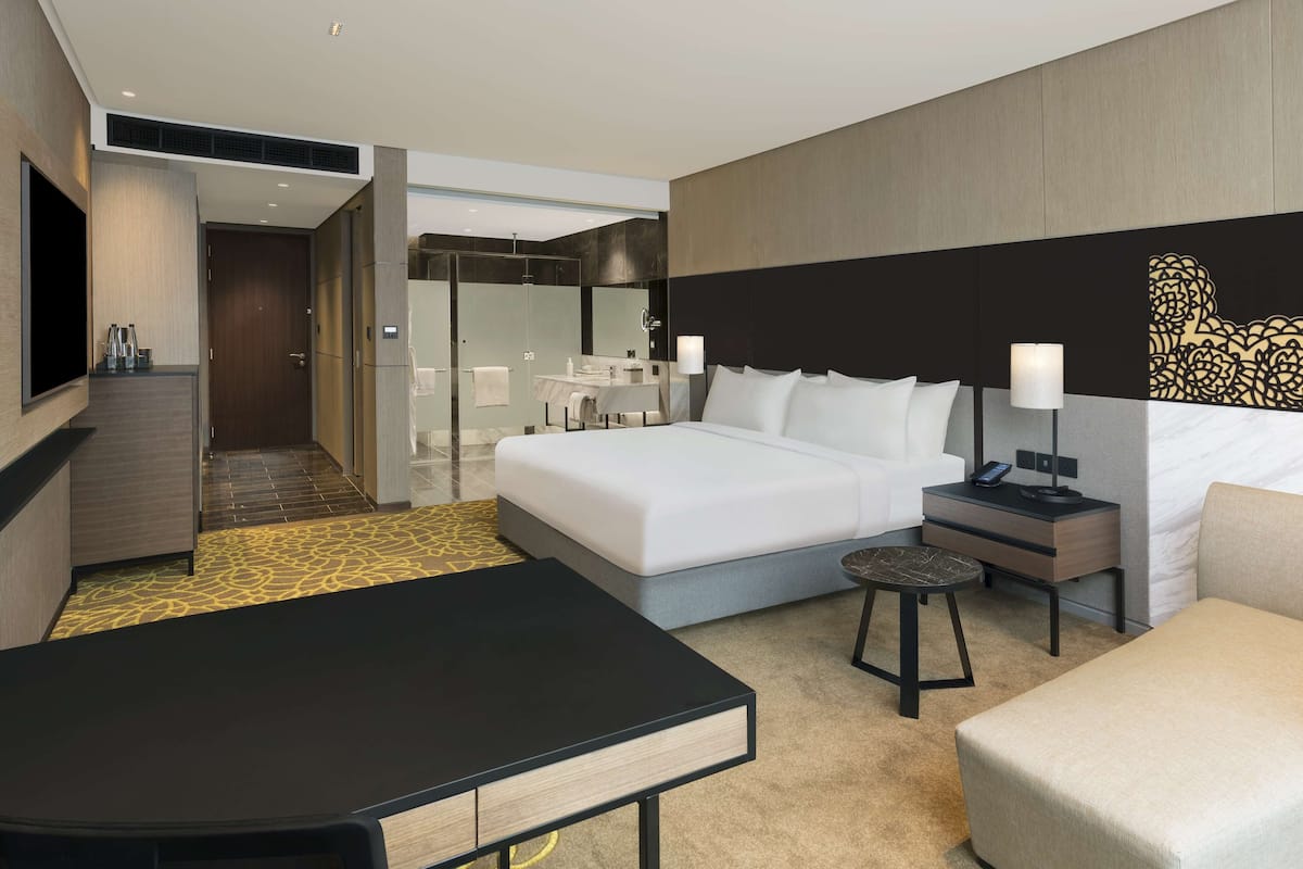 Une chambre d’hôtel moderne équipée d’un grand lit, d’un bureau, d’une chaise, d’un petit tabouret, d’une table de chevet, d’une télévision et d’une salle de bain avec une douche à parois de verre.