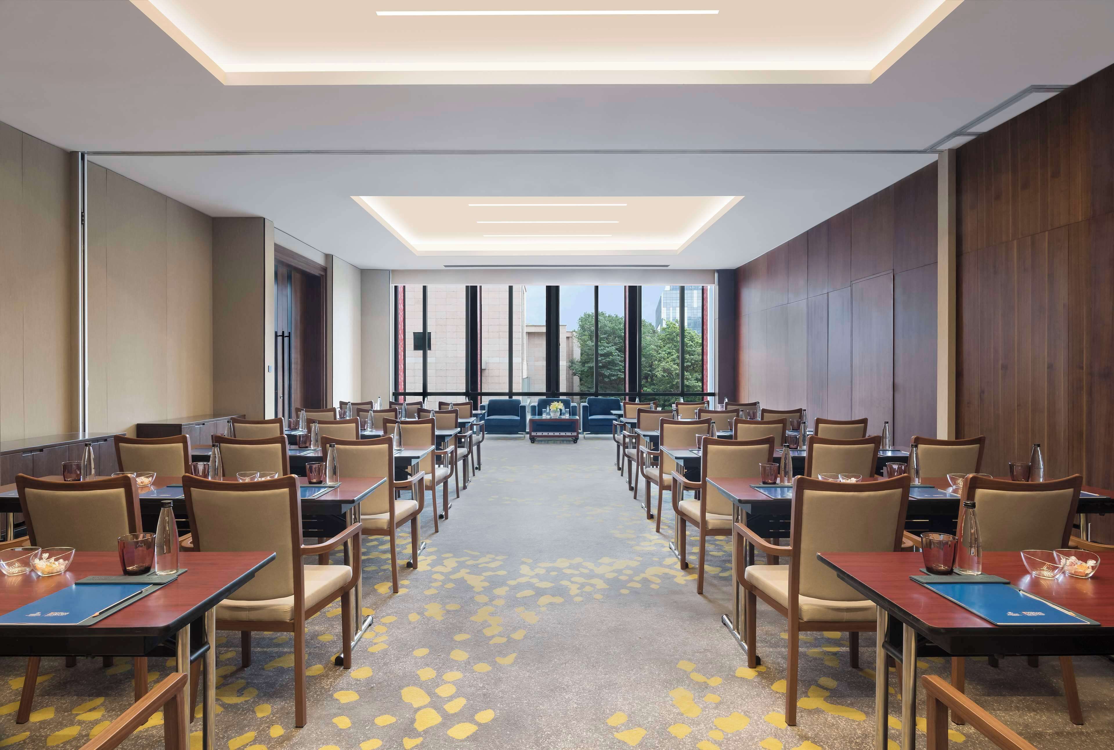 A conference room with rows of tables and chairs, large windows, and a view of buildings and trees.