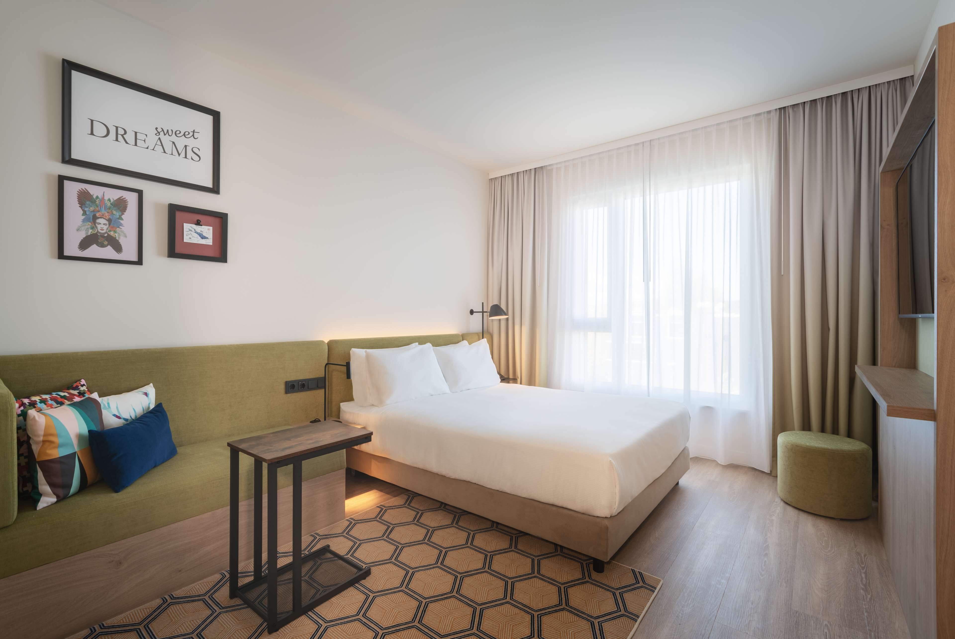 Moderni hotellihuone, jossa on suuri sänky, vihreä sohva, pieni pöytä ja suuri ikkuna, jossa on verhot.