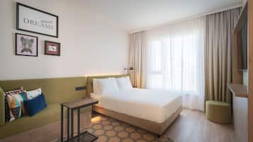 Una habitación de hotel moderna con una cama grande, un sofá verde, una pequeña mesa y un ventanal con cortinas.