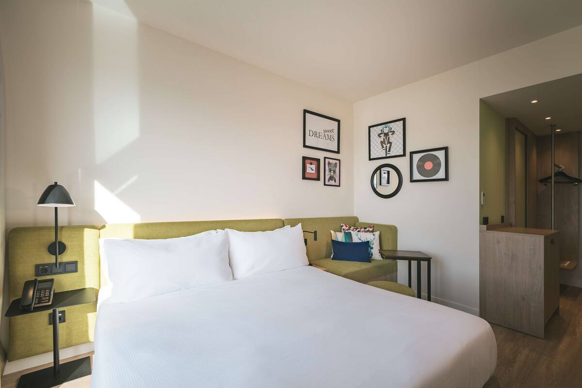 A hotel room with a bed, a bedside table with a lamp, a small desk, and a wardrobe. There are framed pictures on the wall.