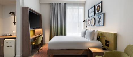 Ein Hotelzimmer mit einem Bett, einem Schreibtisch mit Fernseher, einem Kühlschrank, einem Sessel und einem Nachttisch mit Lampe.