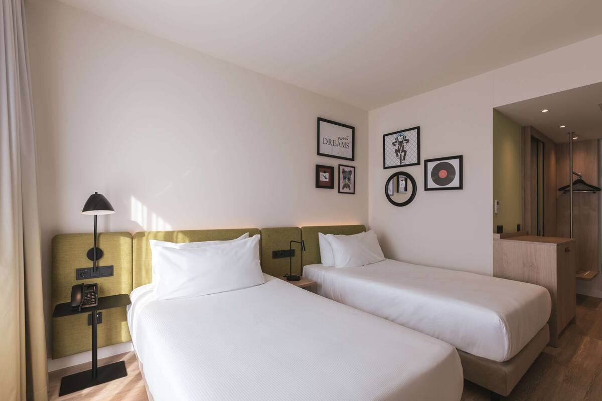 A hotel room with two beds, a bedside table with a lamp, a phone, and framed pictures on the wall.