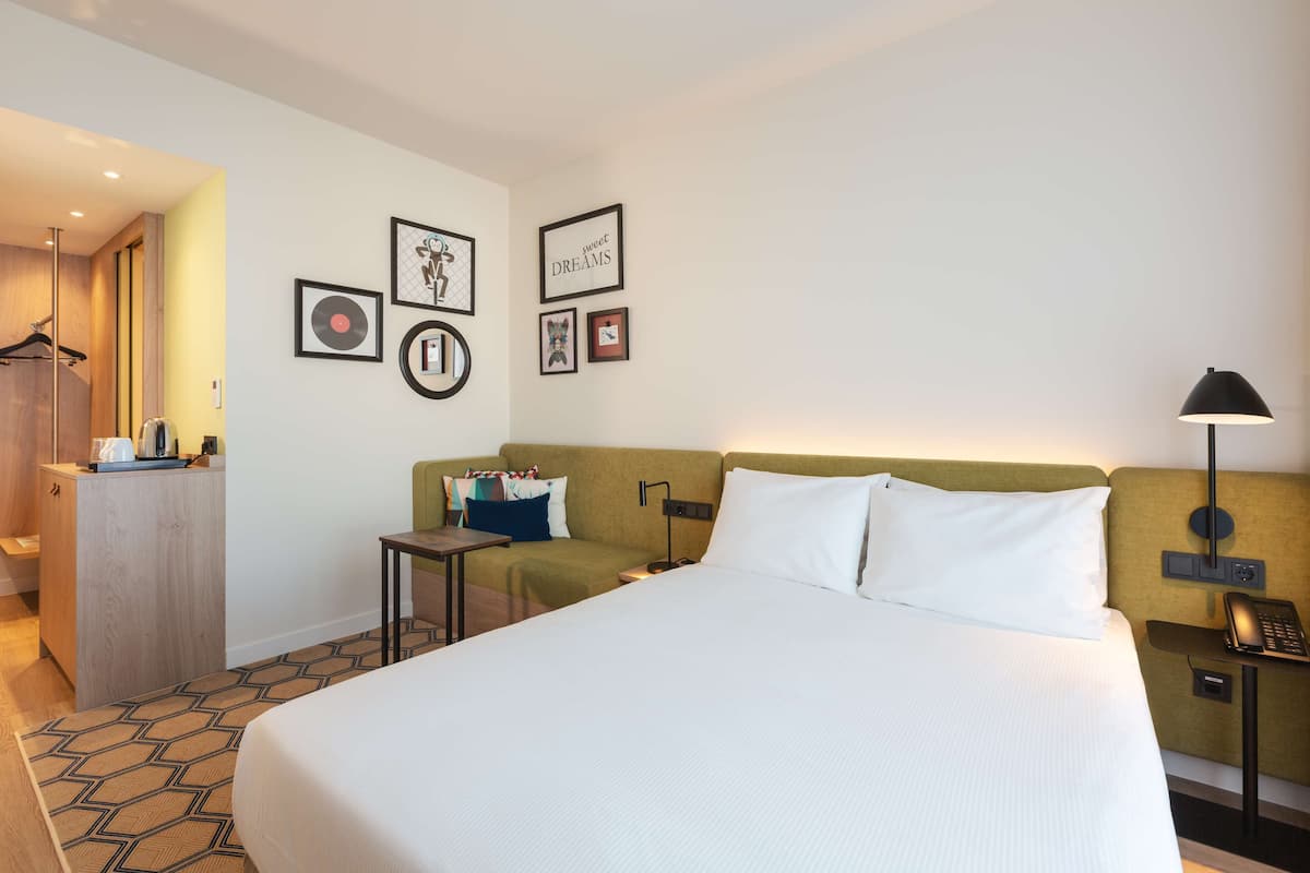A hotel room with a large bed, a desk, a chair, a wardrobe, and a wall with framed pictures.