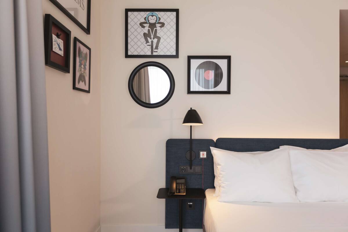 A hotel room with a bed, a nightstand with a lamp, a mirror, and framed artwork on the wall.