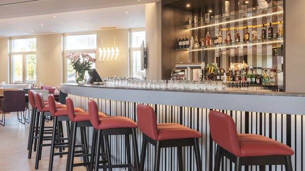 A bar area with a long counter, multiple stools, and a well-stocked shelf of bottles. - Hampton by Hilton Konstanz (Konstanz)