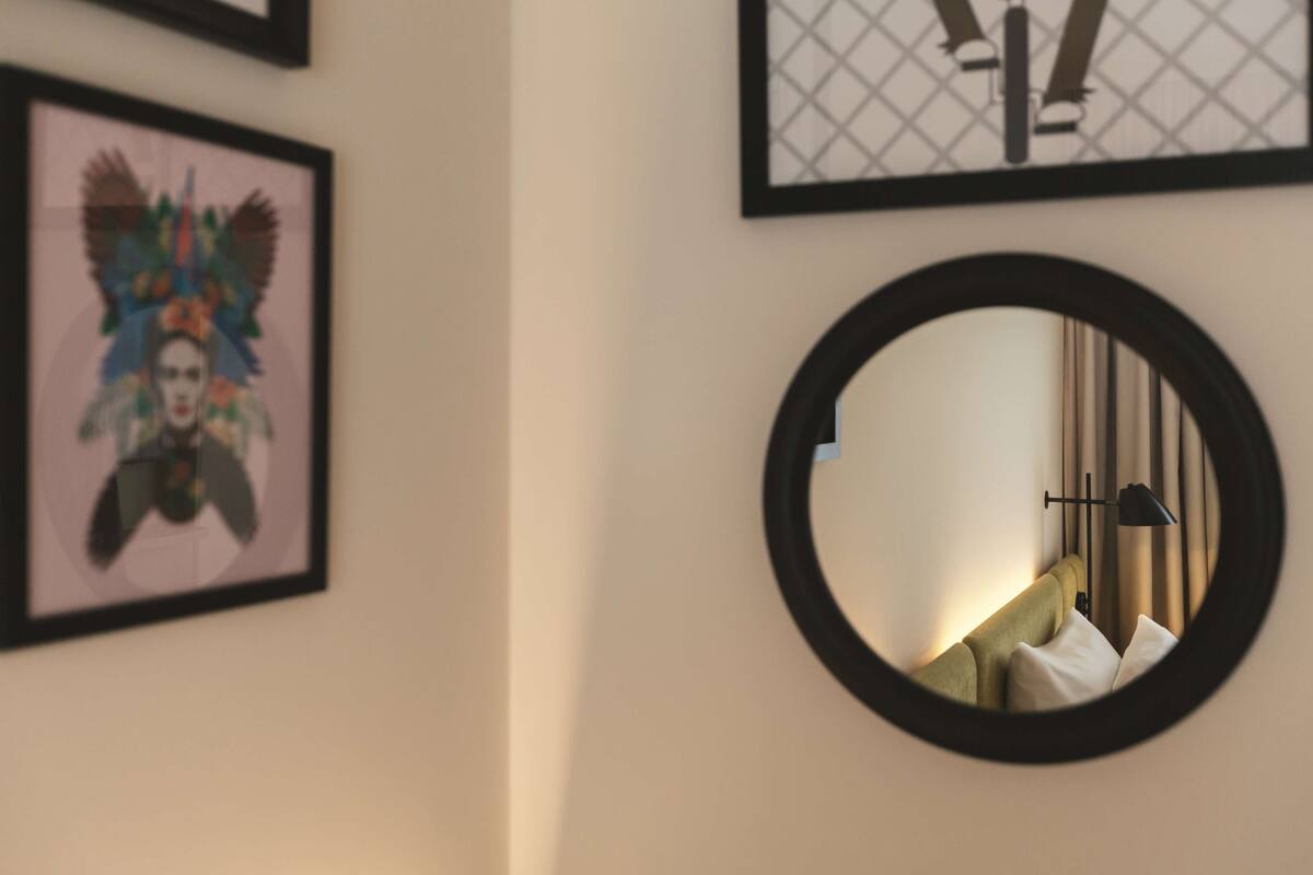 A framed artwork on the wall, a round mirror, and a bed with pillows.