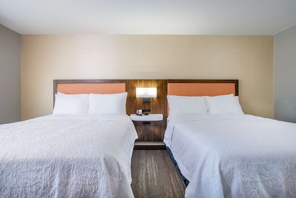 Habitación de hotel con dos camas, cabecera de madera, mesita de noche con lámpara y un lavamanos pequeño.