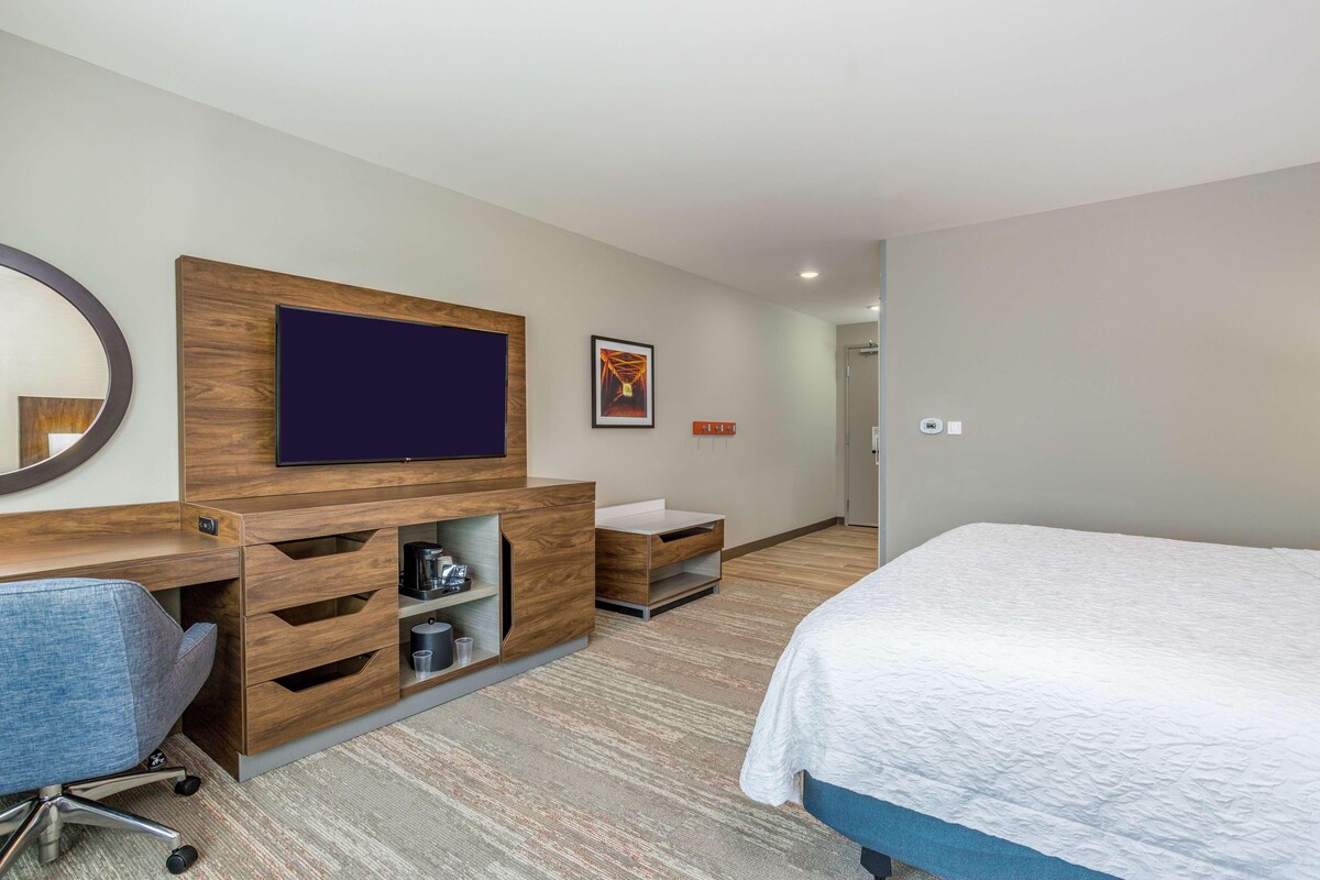 Una habitación de hotel con cama, escritorio con silla, televisión, cafetera y una pequeña estantería.