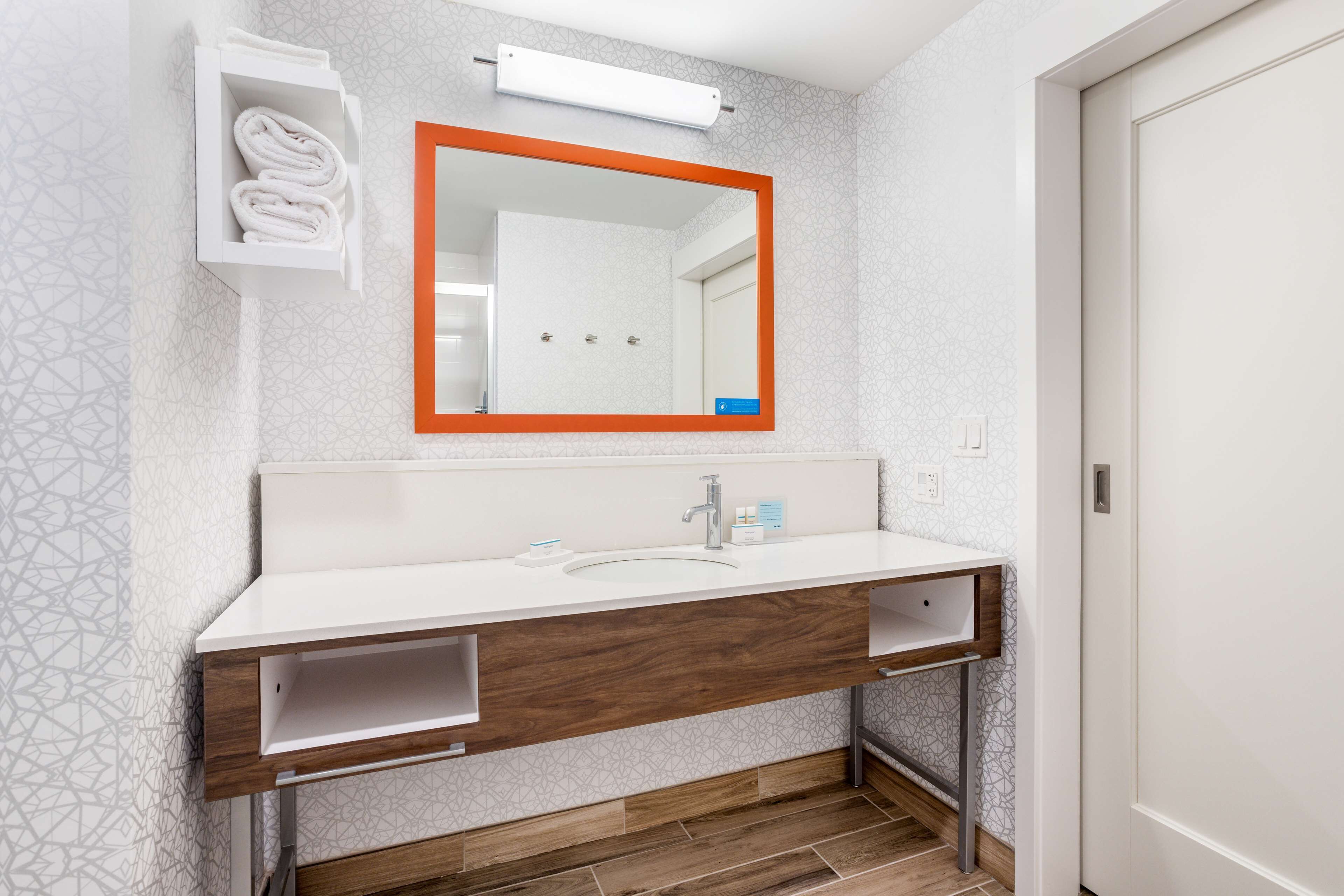 Een moderne badkamer met een witte wastafel, een houten opbergmeubel en een spiegel met oranje rand.